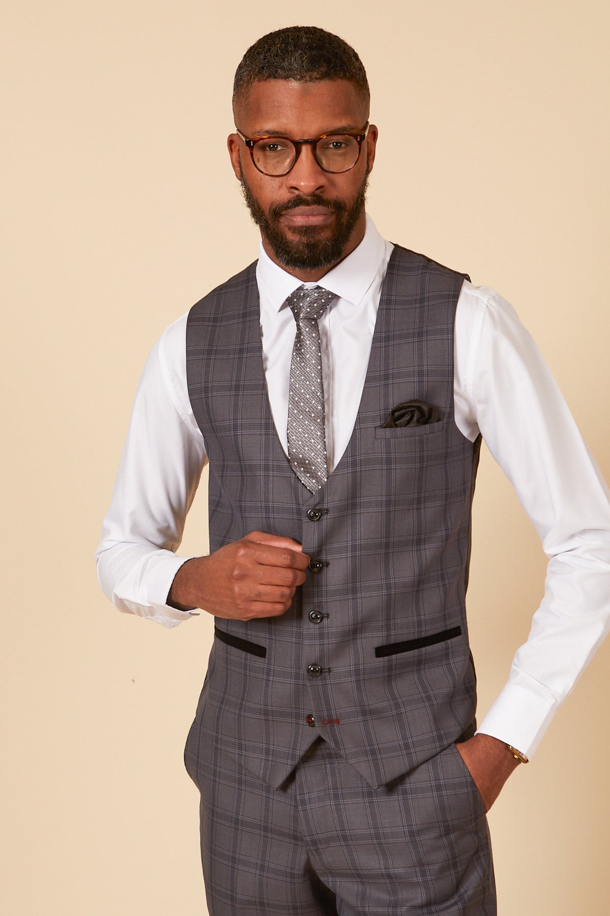 A man with short hair, a beard, and glasses wears the marcdarcy JOSE Grey Check Single Breasted Waistcoat with matching pants over a white shirt, patterned tie, and pocket square, standing against a plain beige background.