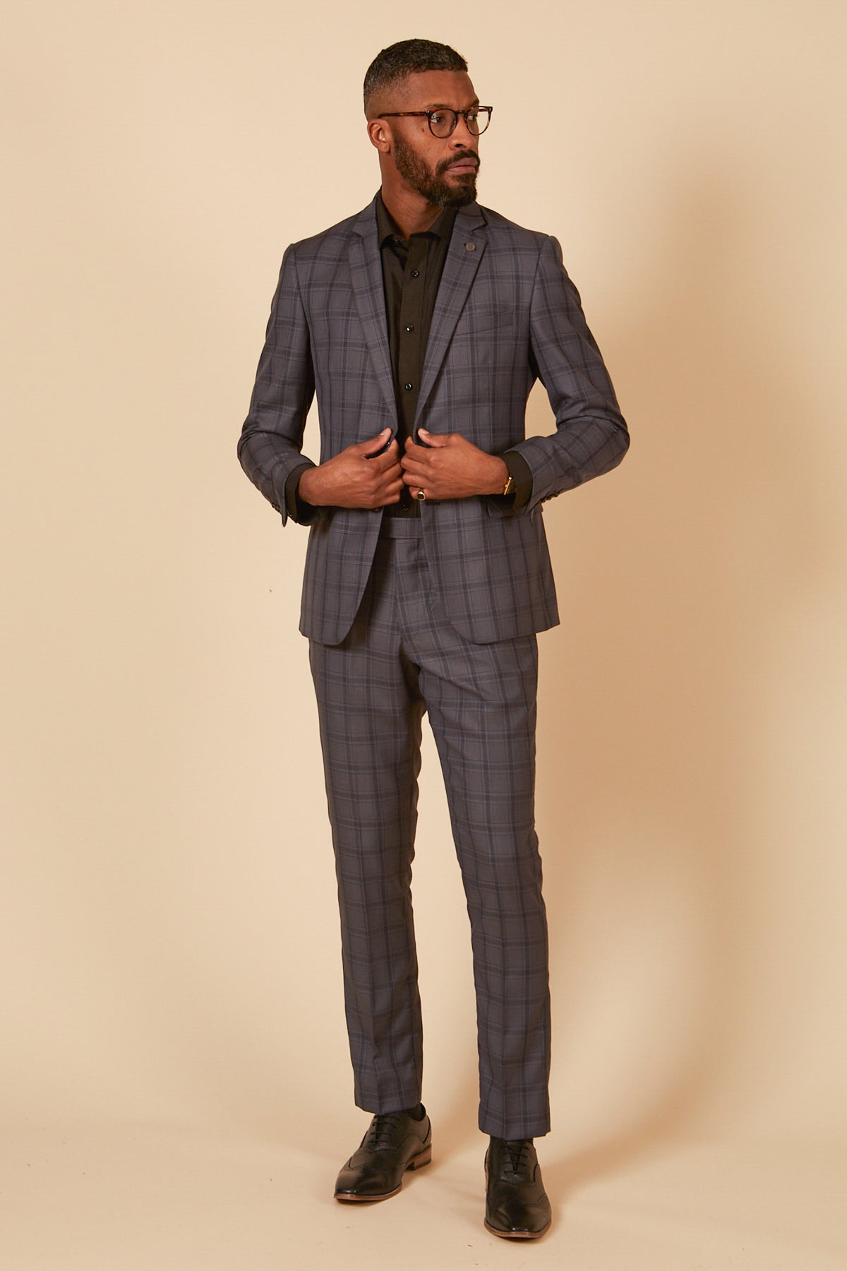 A man wearing glasses and black shoes stands against a beige background, looking to his left while adjusting his tailored JOSE - Grey Check Blazer from Marc Darcy over a black shirt.