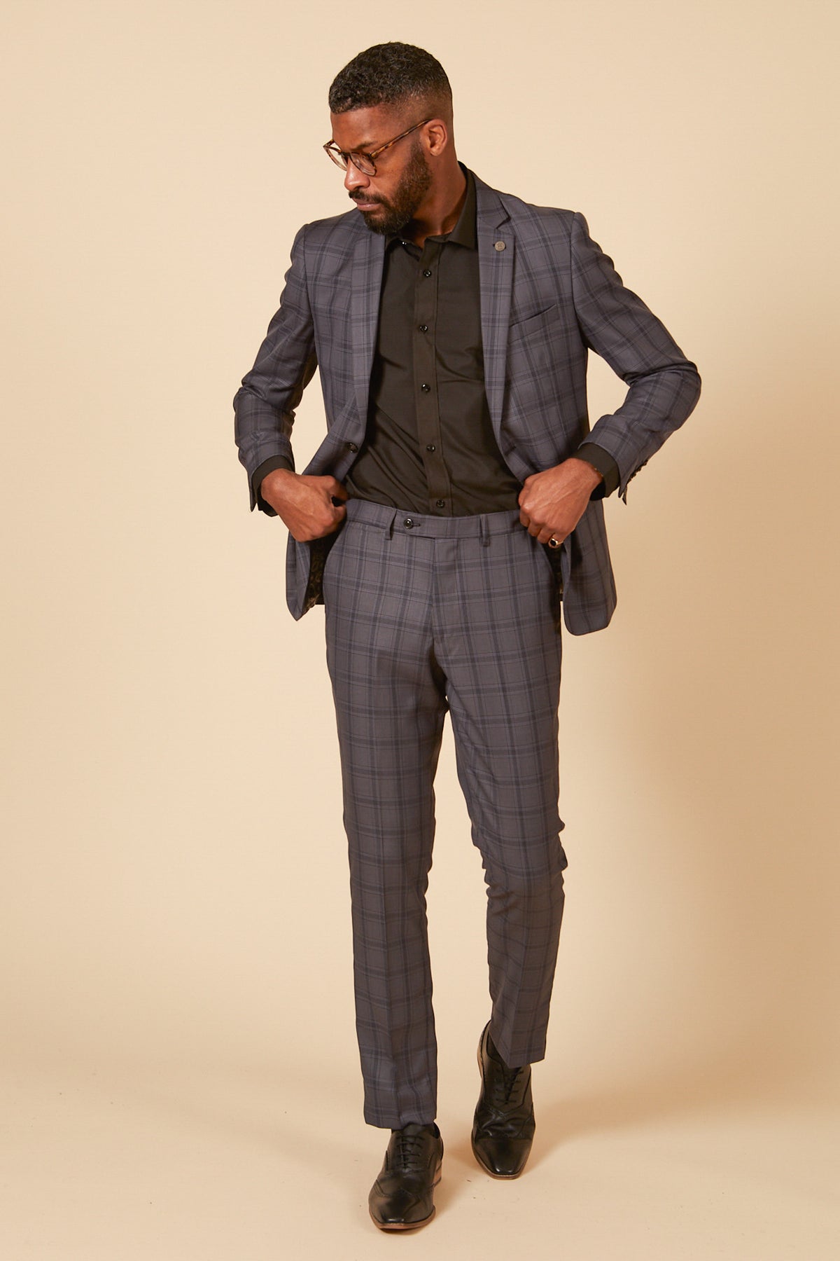 A man in glasses poses against a beige background wearing the Marc Darcy JOSE Grey Check Blazer, paired with a black dress shirt and black shoes, looking down as he adjusts his tailored jacket.