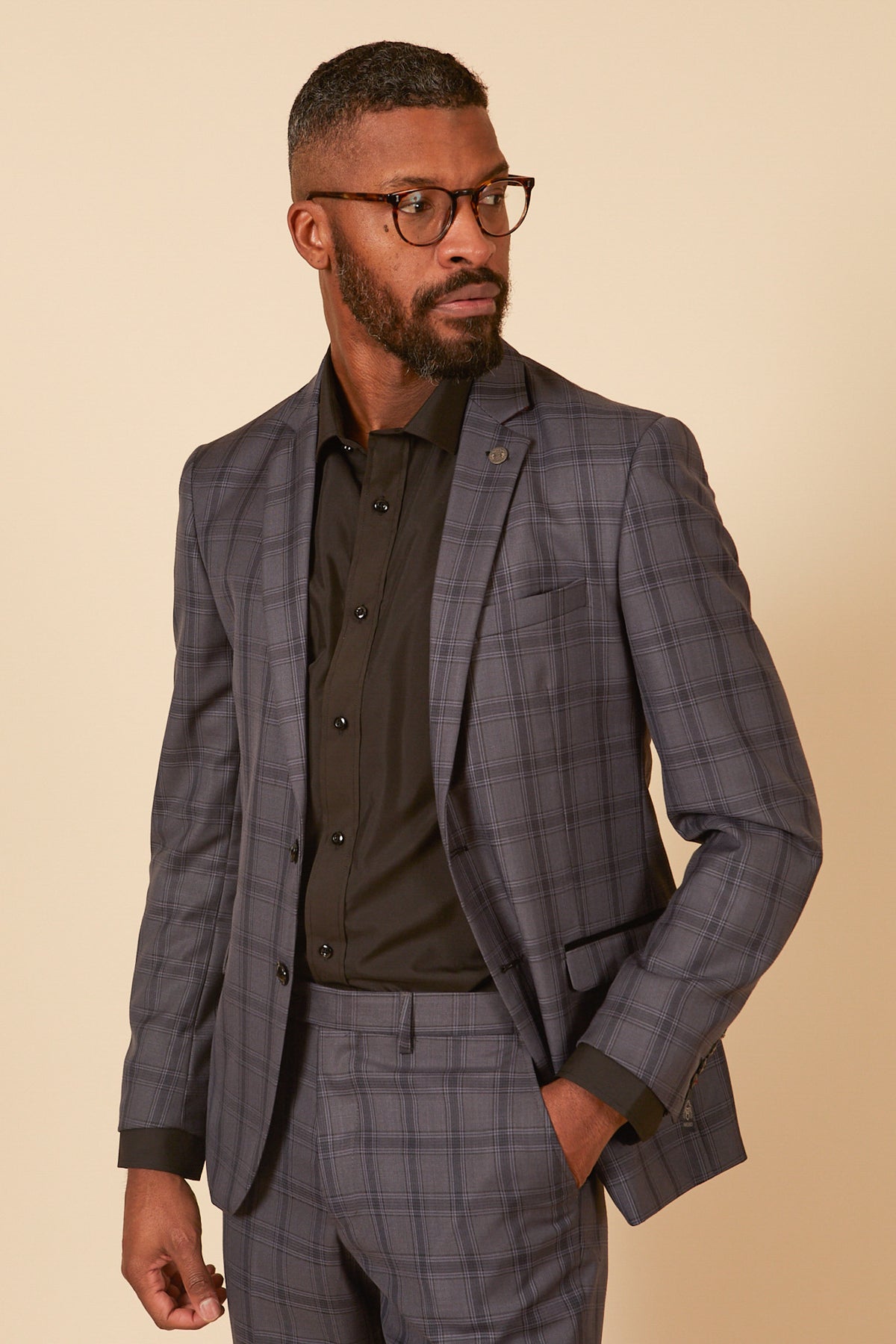 A man with short hair and a beard wears glasses, a black dress shirt, and the JOSE - Grey Check Blazer by Marc Darcy with matching pants. He looks thoughtfully to the side against a neutral background.