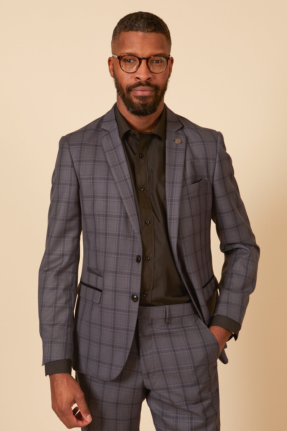 A man with short hair, a beard, and glasses wears the Marc Darcy JOSE Grey Check Blazer over a black dress shirt. He stands against a light background, one hand in his pocket and a slight smile on his face.