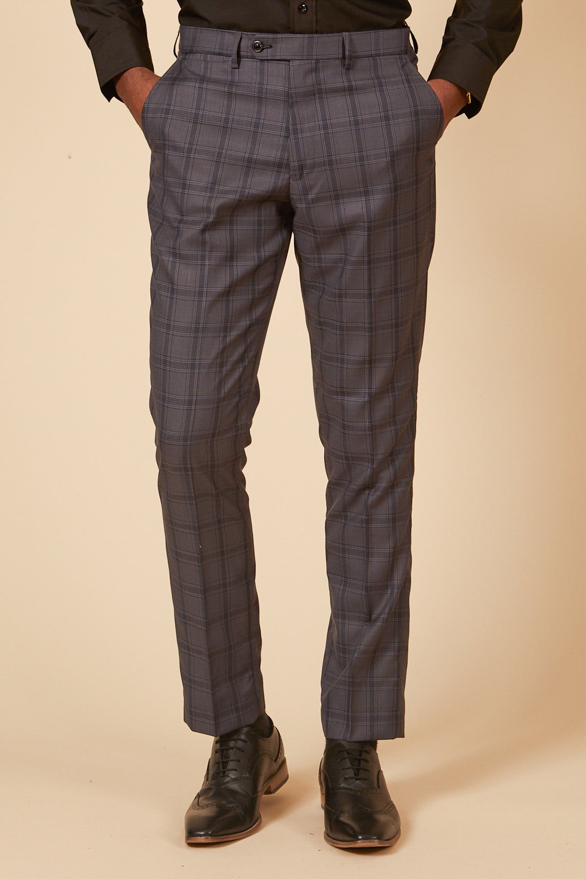 Man dressed in the marcdarcy JOSE Grey Check Three Piece Suit pants, paired with a black shirt and black brogue shoes, stands with his hands in his pockets.
