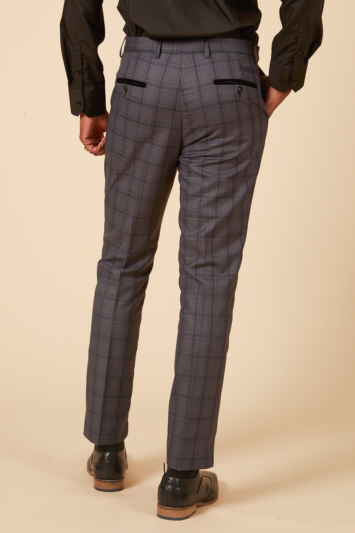 A man wears marcdarcy JOSE grey check trousers with a black shirt and black dress shoes, set against a cream background.
