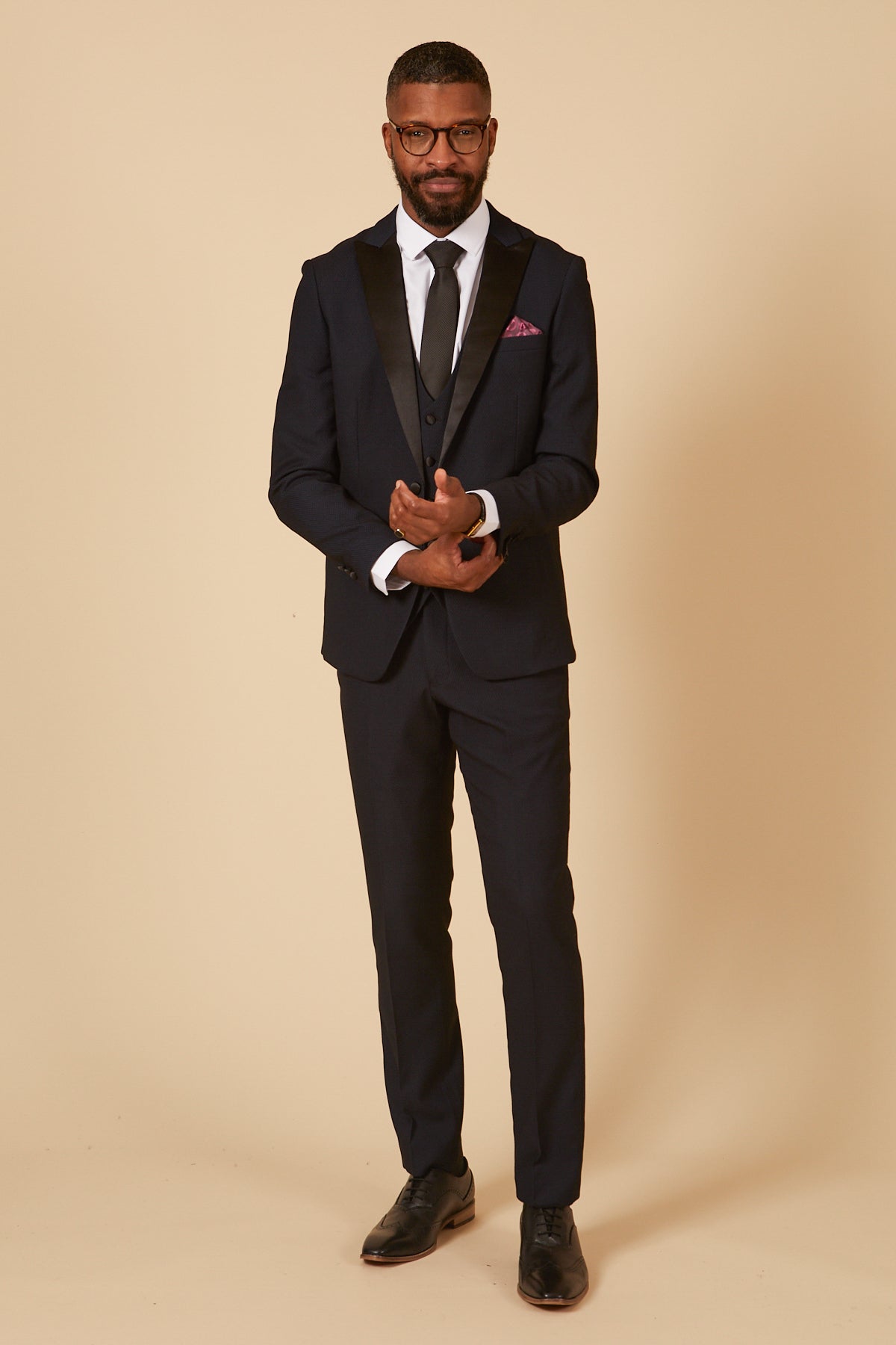 A man wearing glasses and the marcdarcy DALTON - Navy Tux Lapel Diamond Three Piece Suit stands against a beige background, adjusting his cuff with a slight smile.