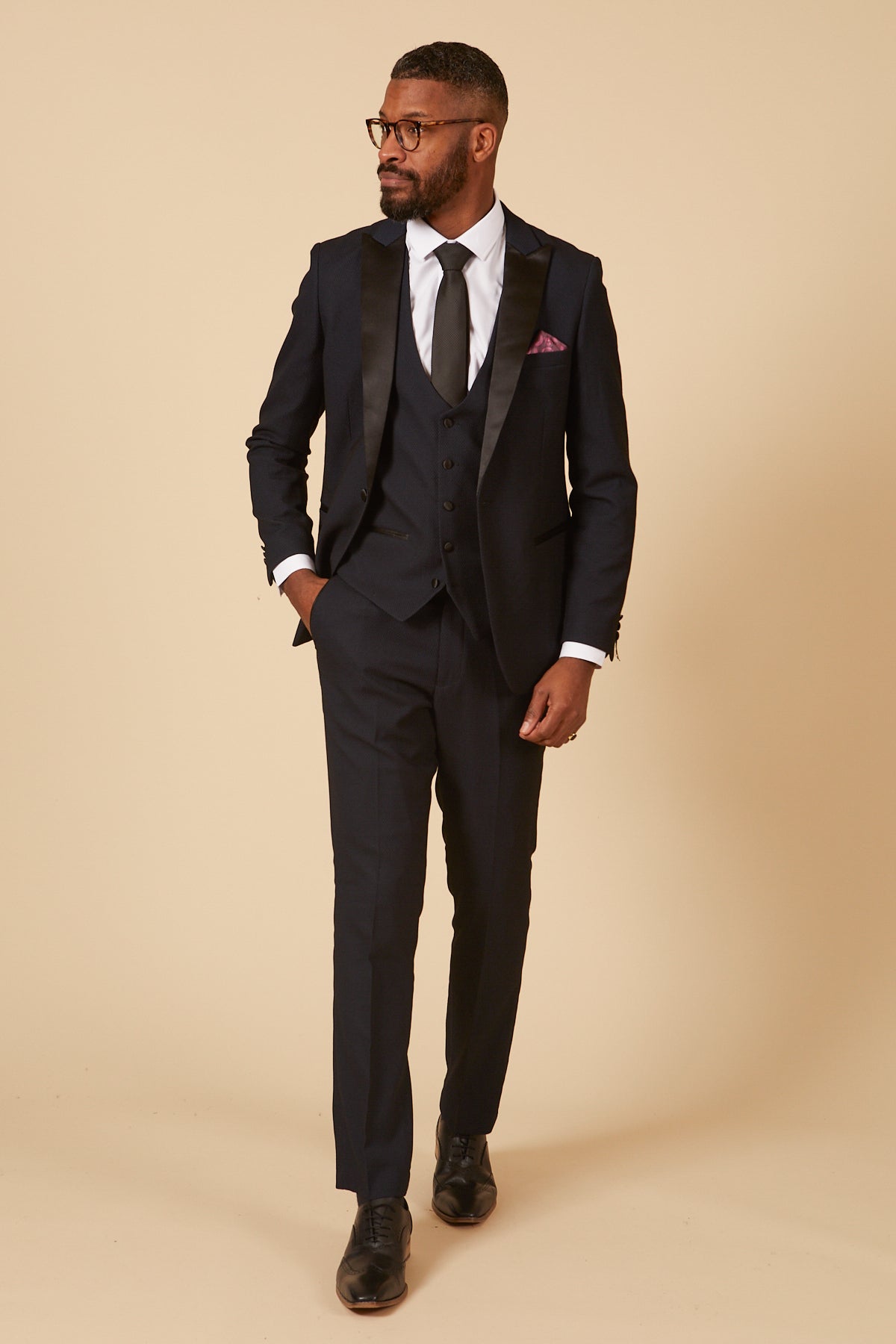 A man wearing the marcdarcy DALTON Navy Tux Lapel Diamond Three Piece Suit with black satin lapels, white shirt, black tie, matching vest, slim fit, black shoes, and a purple pocket square stands against a beige background.