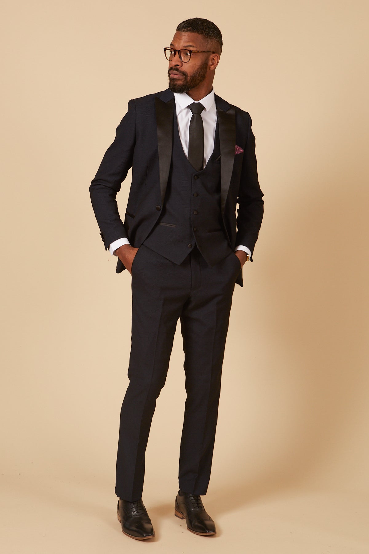 A man wearing the marcdarcy DALTON Navy Tux Lapel Diamond Three Piece Suit, paired with a white shirt, black tie, and glasses stands with his hands in his pockets against a cream background.
