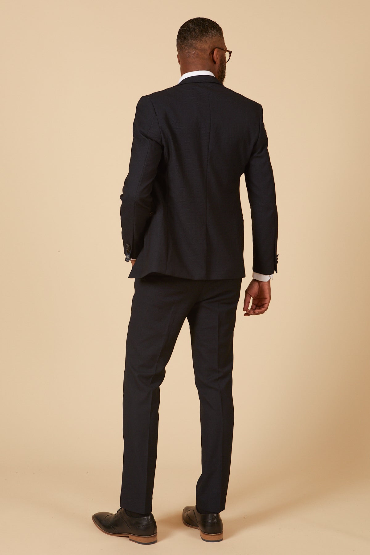 A man stands with his back to the camera against a beige background, wearing the marcdarcy DALTON Navy Tux Lapel Diamond Three Piece Suit.