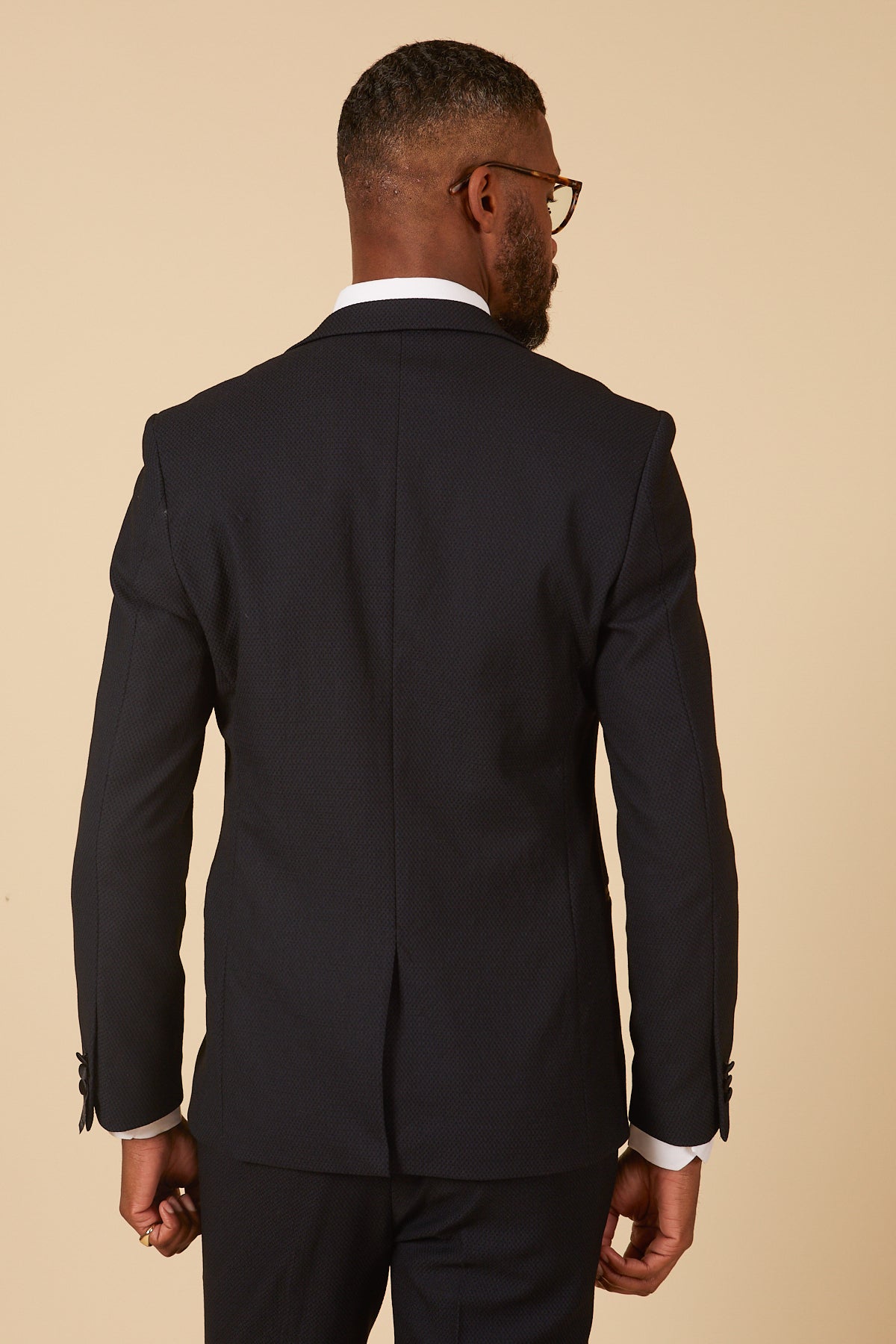 A man with short hair and glasses is seen from behind, wearing the marcdarcy DALTON - Navy Tux Lapel Diamond Blazer over a white dress shirt, set against a plain beige background.