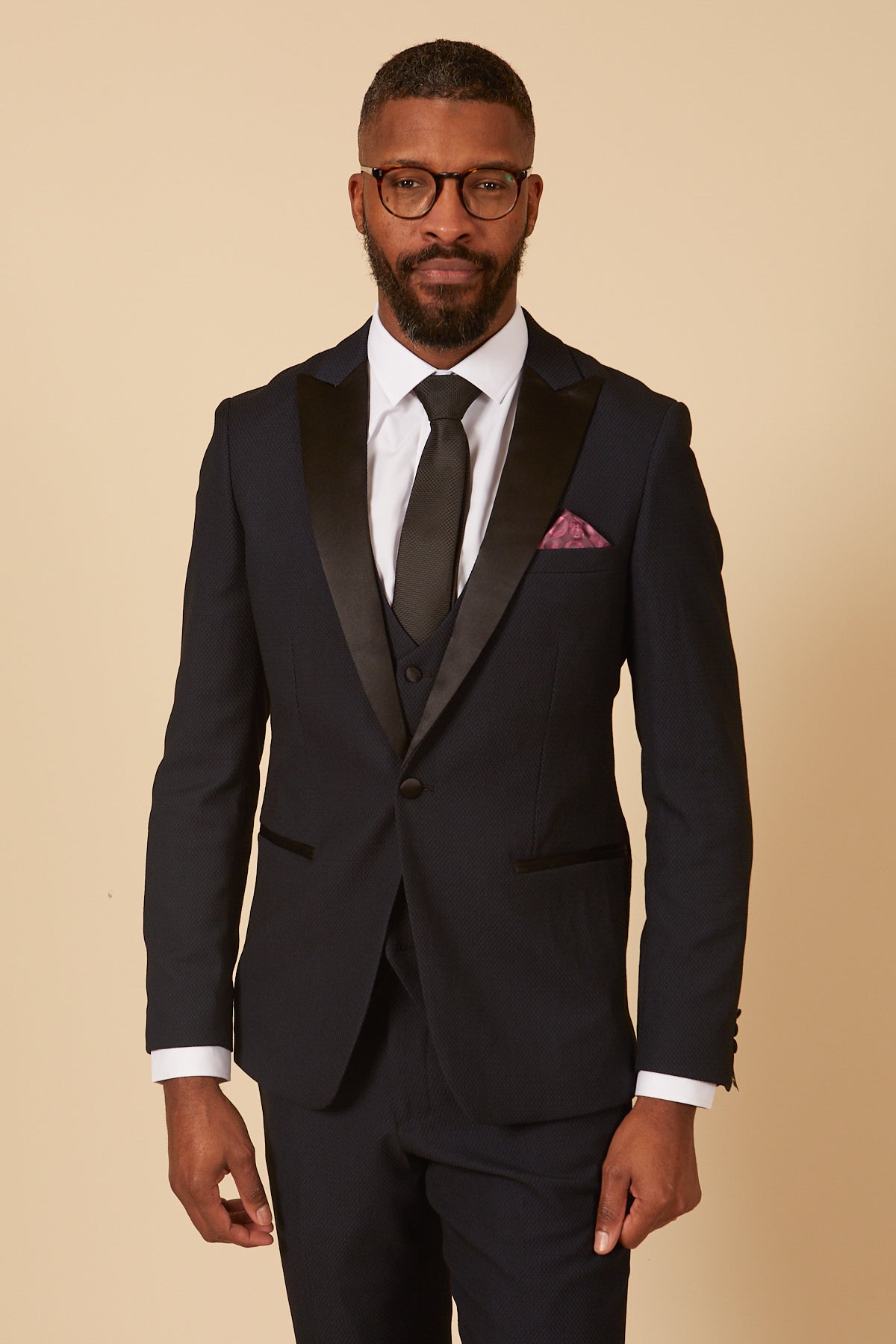 A man with short hair, a beard, and glasses wears the marcdarcy DALTON Navy Tux Lapel Diamond Three Piece Suit with a black satin peak lapel, white shirt, black tie, and purple pocket square against a beige background.