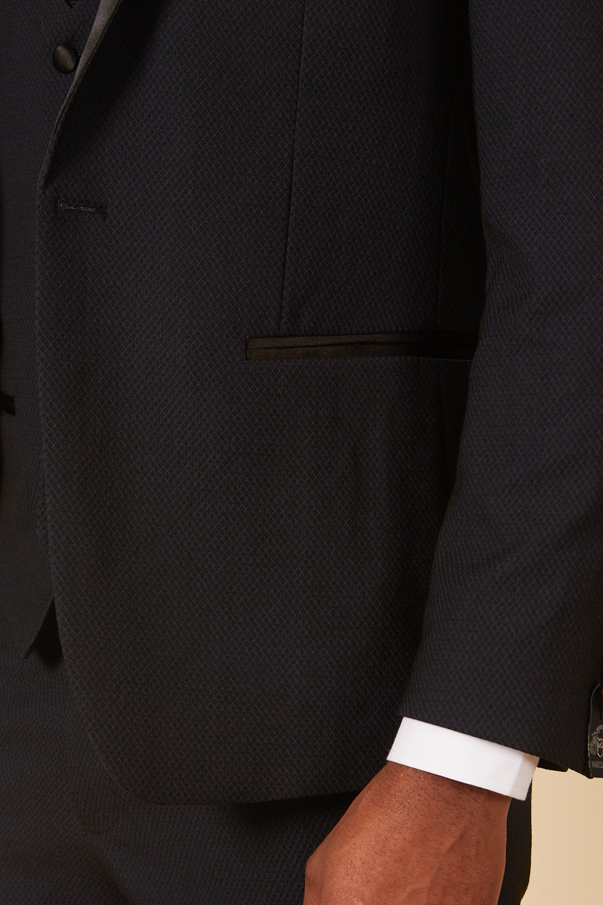 A close-up of a person wearing the marcdarcy DALTON - Navy Tux Lapel Diamond Blazer featuring a textured navy suit jacket with black-trimmed pocket, white shirt cuff visible, and their hand resting by their side.
