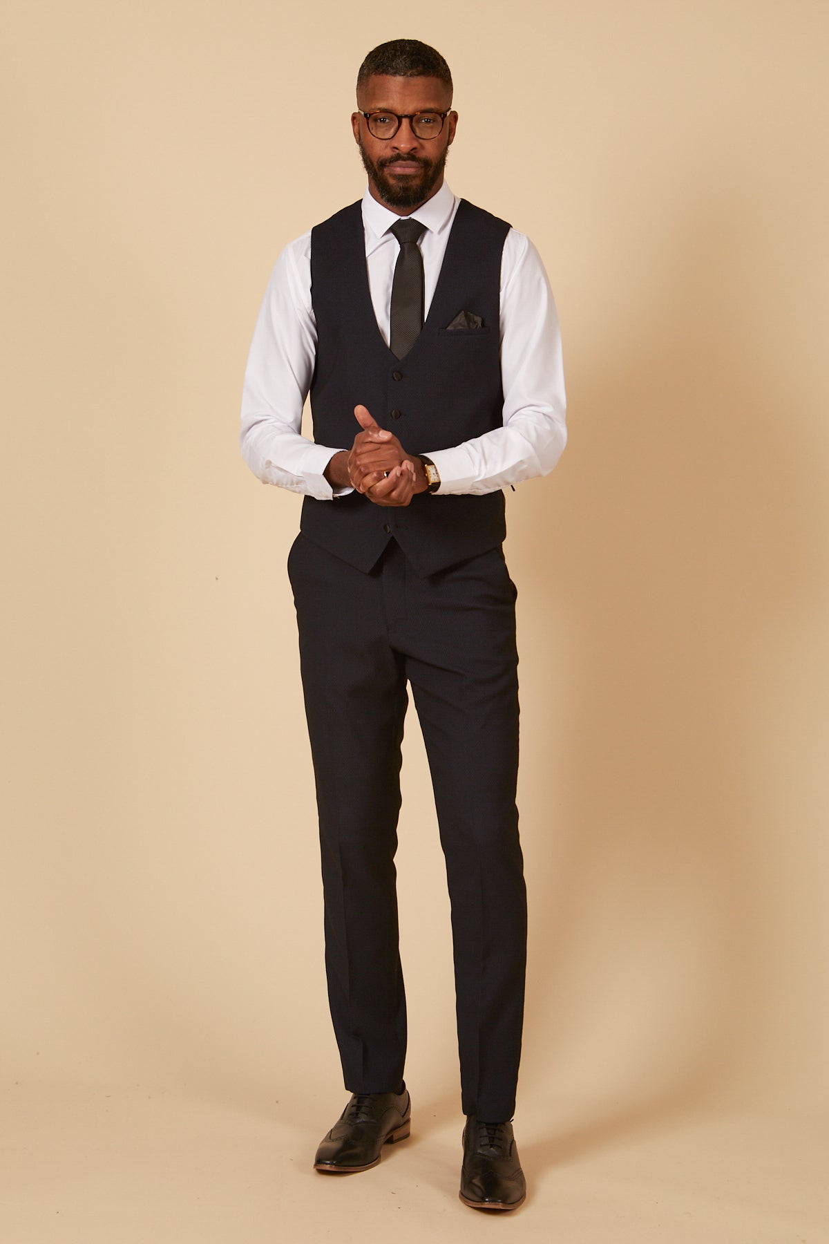 A man in glasses wears the marcdarcy DALTON - Navy Diamond Waistcoat with matching trousers, a white dress shirt, black tie, and black dress shoes, standing against a beige background and looking at the camera with hands clasped.