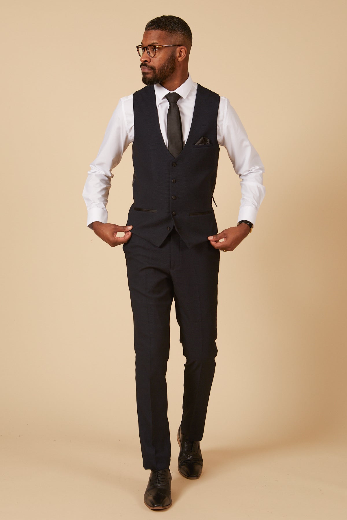 A man in glasses and a white dress shirt wears the marcdarcy DALTON Navy Tux Lapel Diamond Three Piece Suit—featuring a slim-fit dark vest, matching trousers, black tie, and shoes—standing against a beige background adjusting his vest.