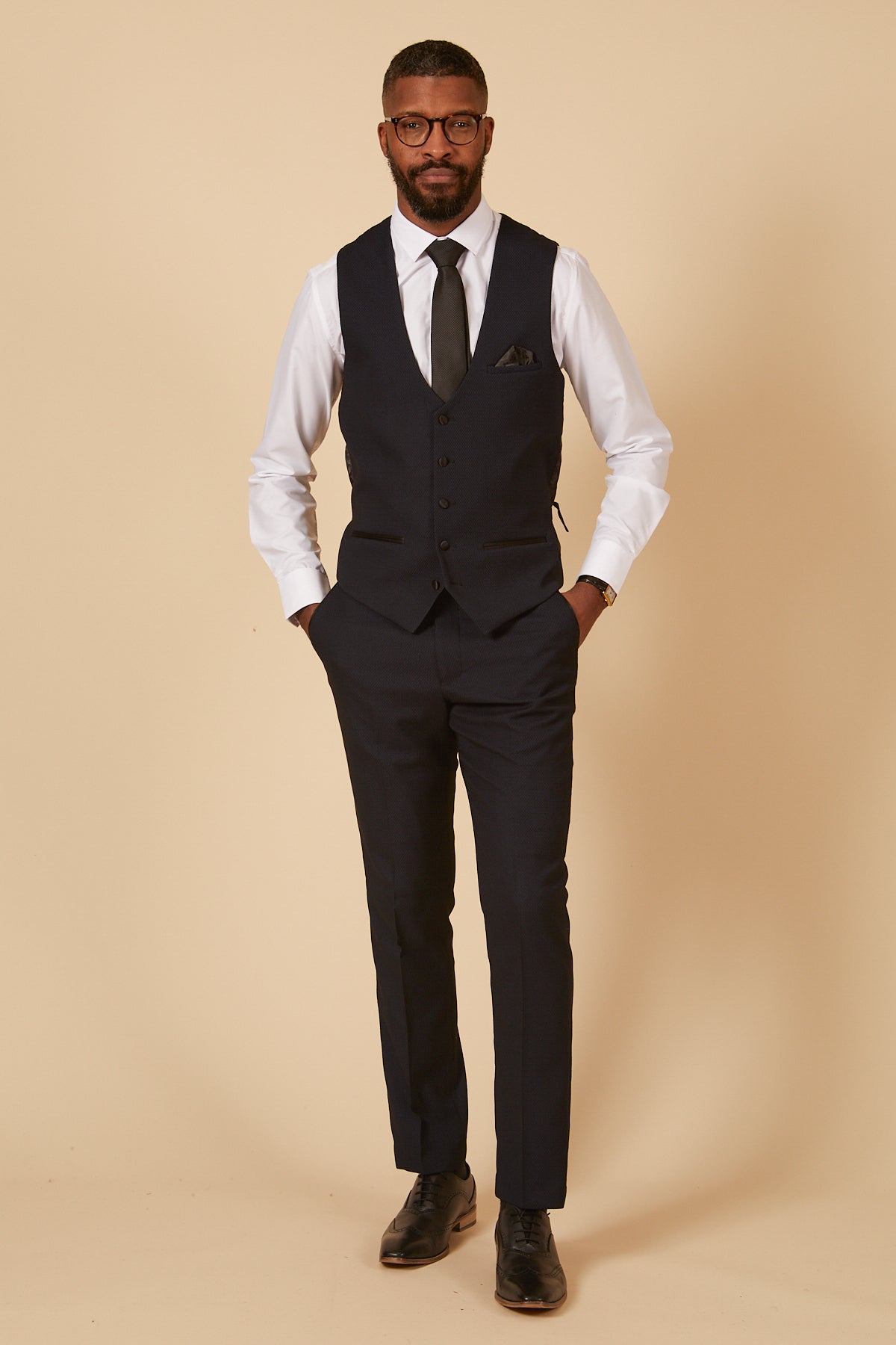 A man wears the marcdarcy DALTON - Navy Tux Lapel Diamond Three Piece Suit with a waistcoat, white shirt, black tie, pocket square, trousers, black Oxford shoes, and glasses, posing against a beige background.