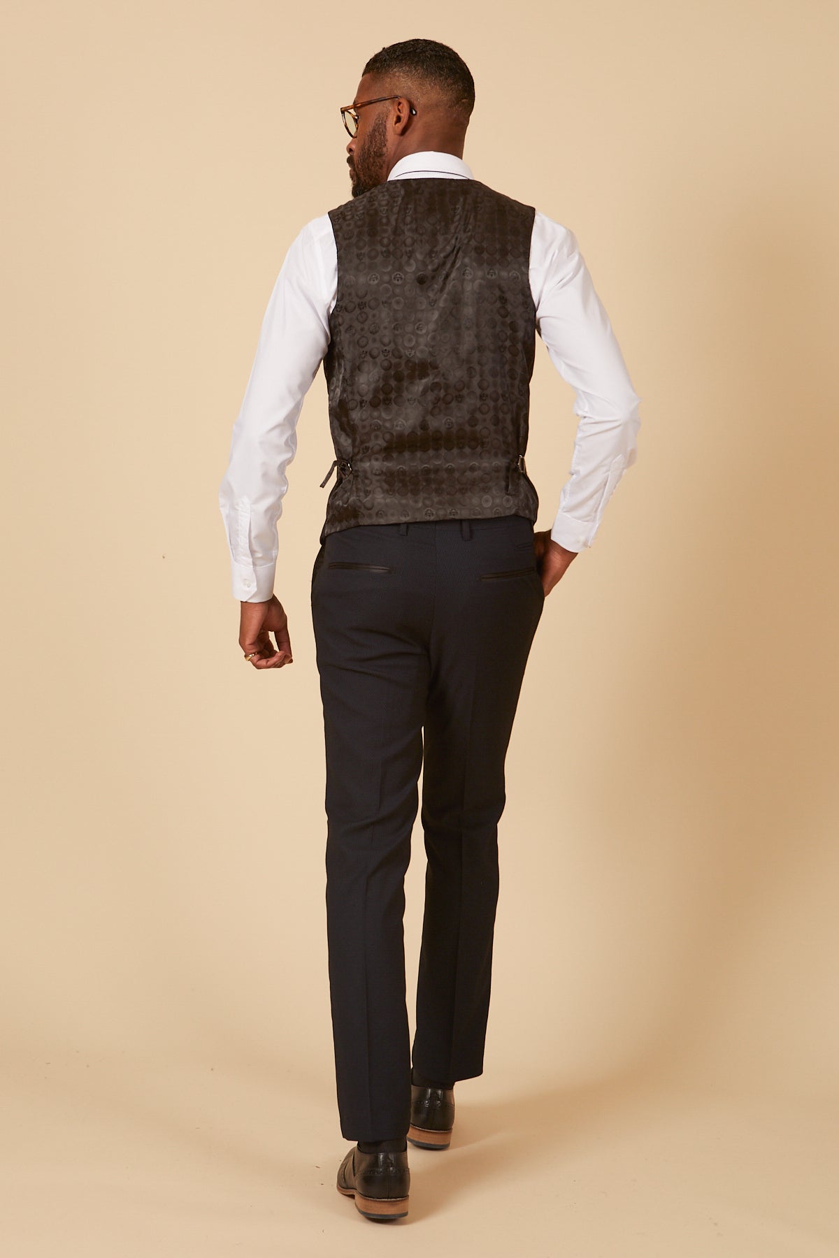 Rear view of a Black man in a white dress shirt, marcdarcy DALTON Navy Diamond Waistcoat, and navy trousers. He has one hand in his pocket, wears glasses and dress shoes.