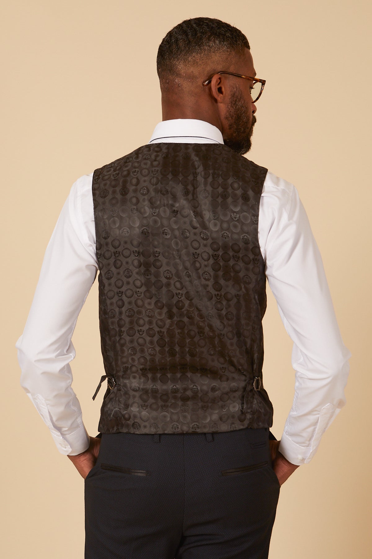 A man faces away from the camera wearing the marcdarcy DALTON - Navy Diamond Waistcoat over a white long-sleeve shirt and dark trousers, standing against a beige background.