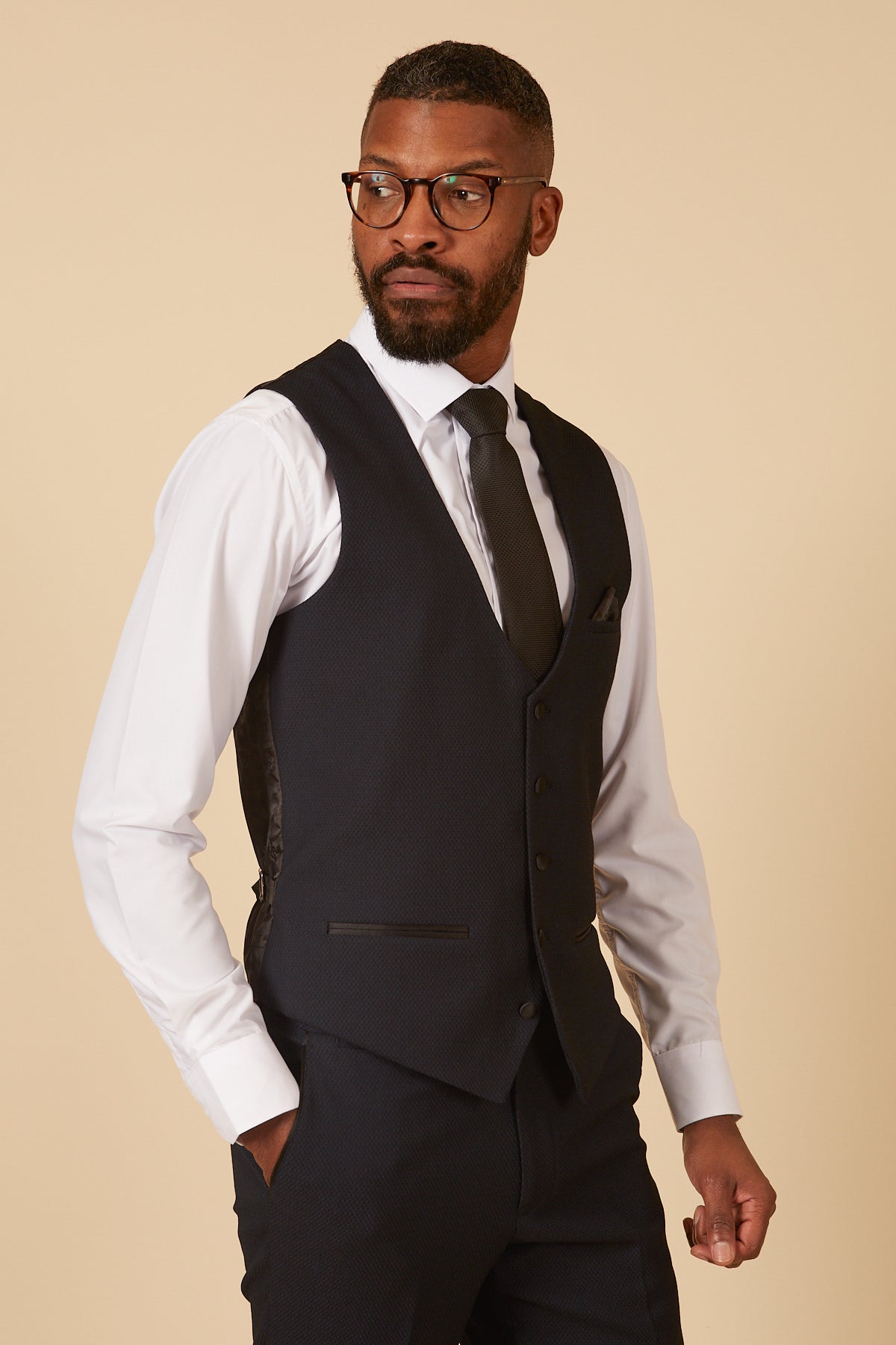 A Black man wearing the marcdarcy DALTON Navy Diamond Waistcoat over a white dress shirt, black tie, and glasses, with his hand in his pocket.