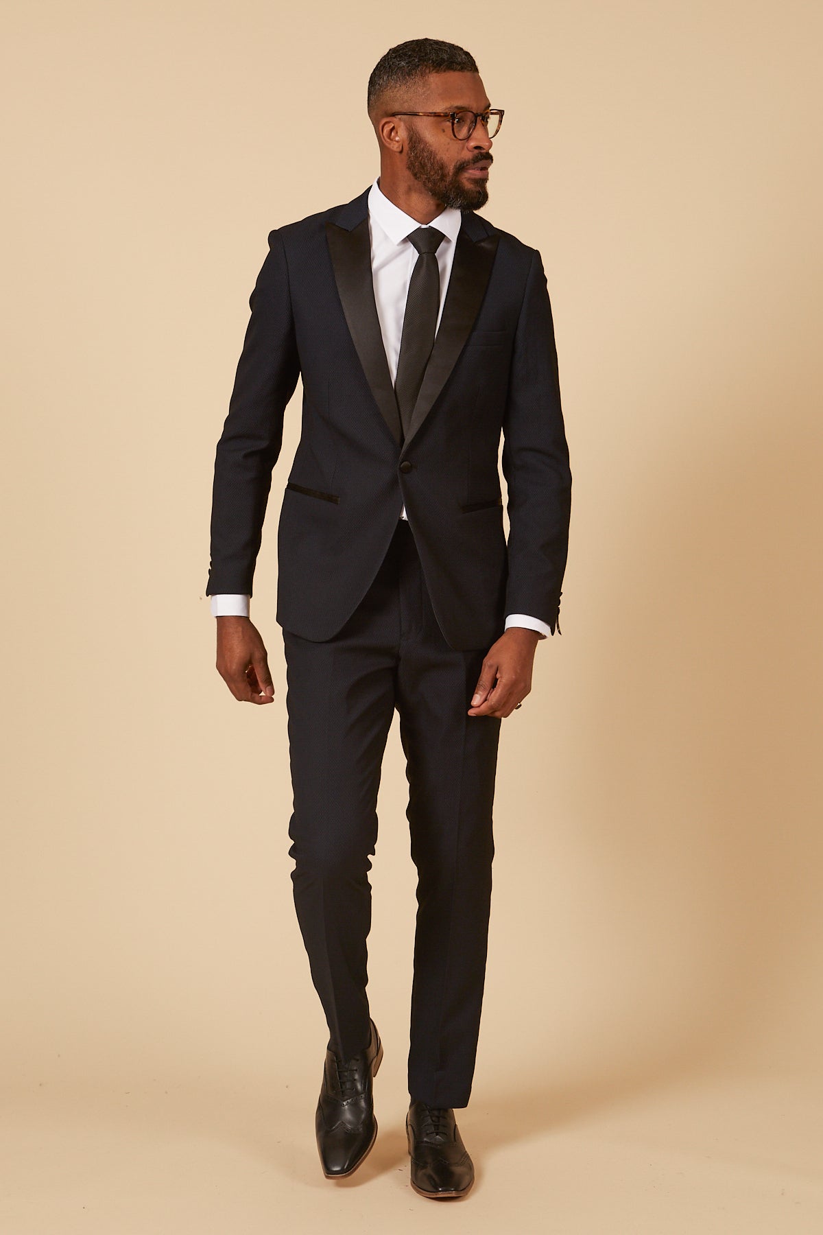 A man with short hair, beard, and glasses wears the marcdarcy DALTON - Navy Tux Lapel Diamond Blazer with a white shirt, black tie, and shoes, standing against a beige background and looking to his left.
