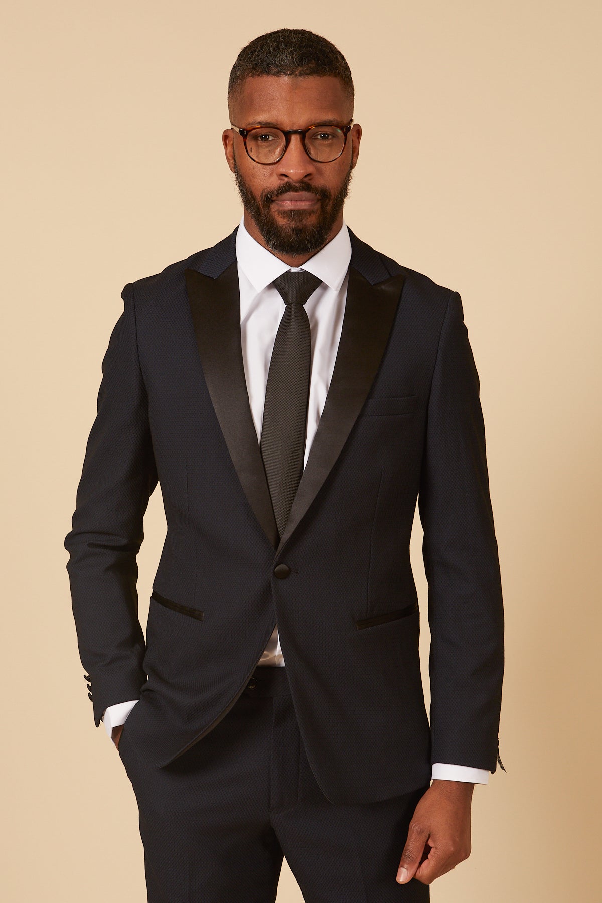 A man with short hair, a beard, and glasses wears the marcdarcy DALTON Navy Tux Lapel Diamond Blazer, paired with a white shirt and black tie, standing against a beige background—perfect for formal events.