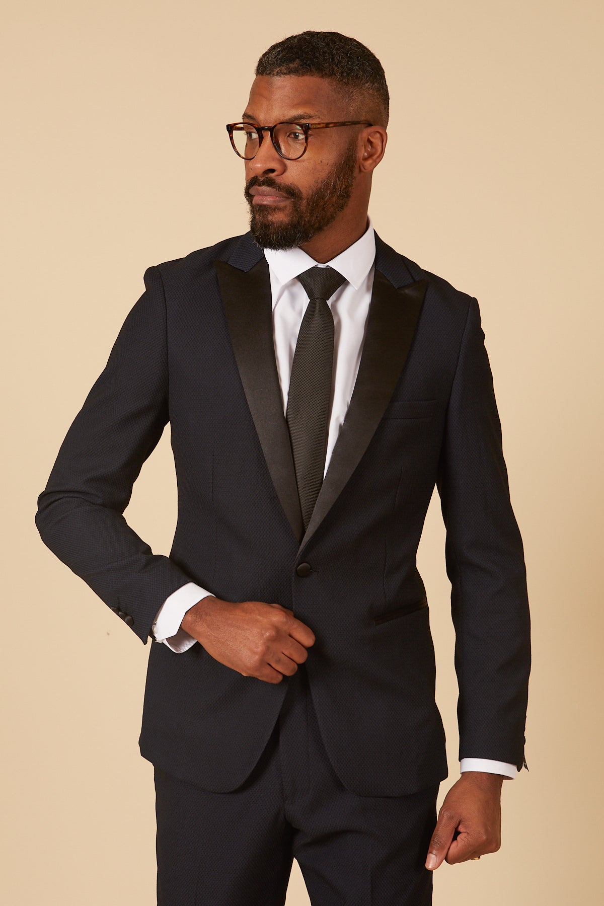 A man in glasses wears the marcdarcy DALTON Navy Tux Lapel Diamond Blazer with a white dress shirt and black tie, posing against a beige background while adjusting his jacket and looking to the side.