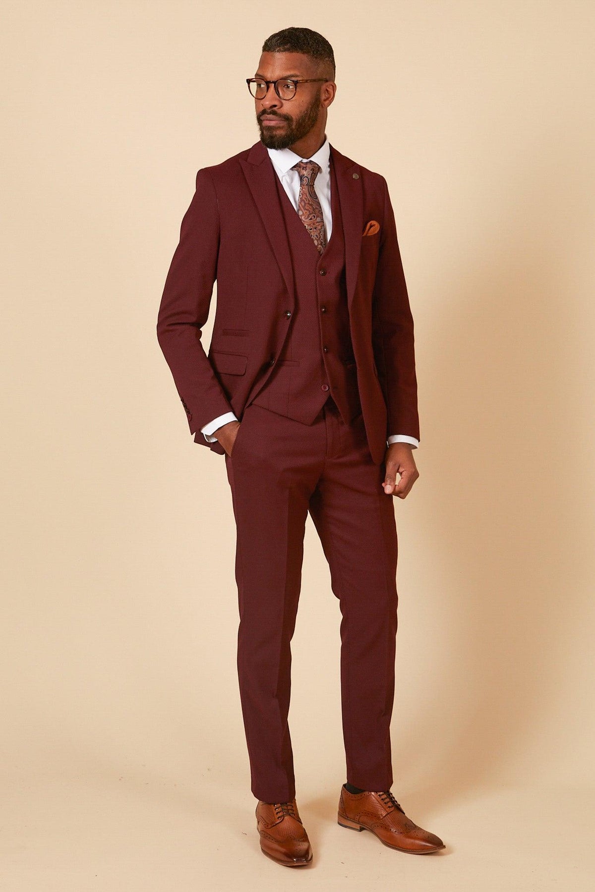 A man in the marcdarcy MAX - Wine Three Piece Suit, paired with a white shirt, patterned tie, glasses, and brown shoes stands against a beige background—a sophisticated option for a wedding guest.