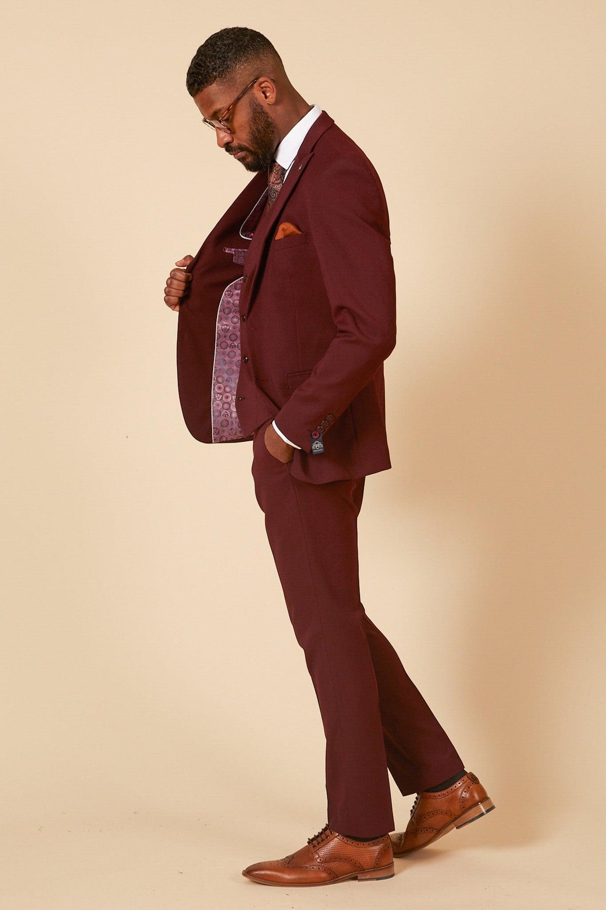 A man in the marcdarcy MAX - Wine Three Piece Suit with a white shirt, brown dress shoes, and an orange pocket square stands sideways, holding open his jacket to reveal its patterned lining against a beige background.