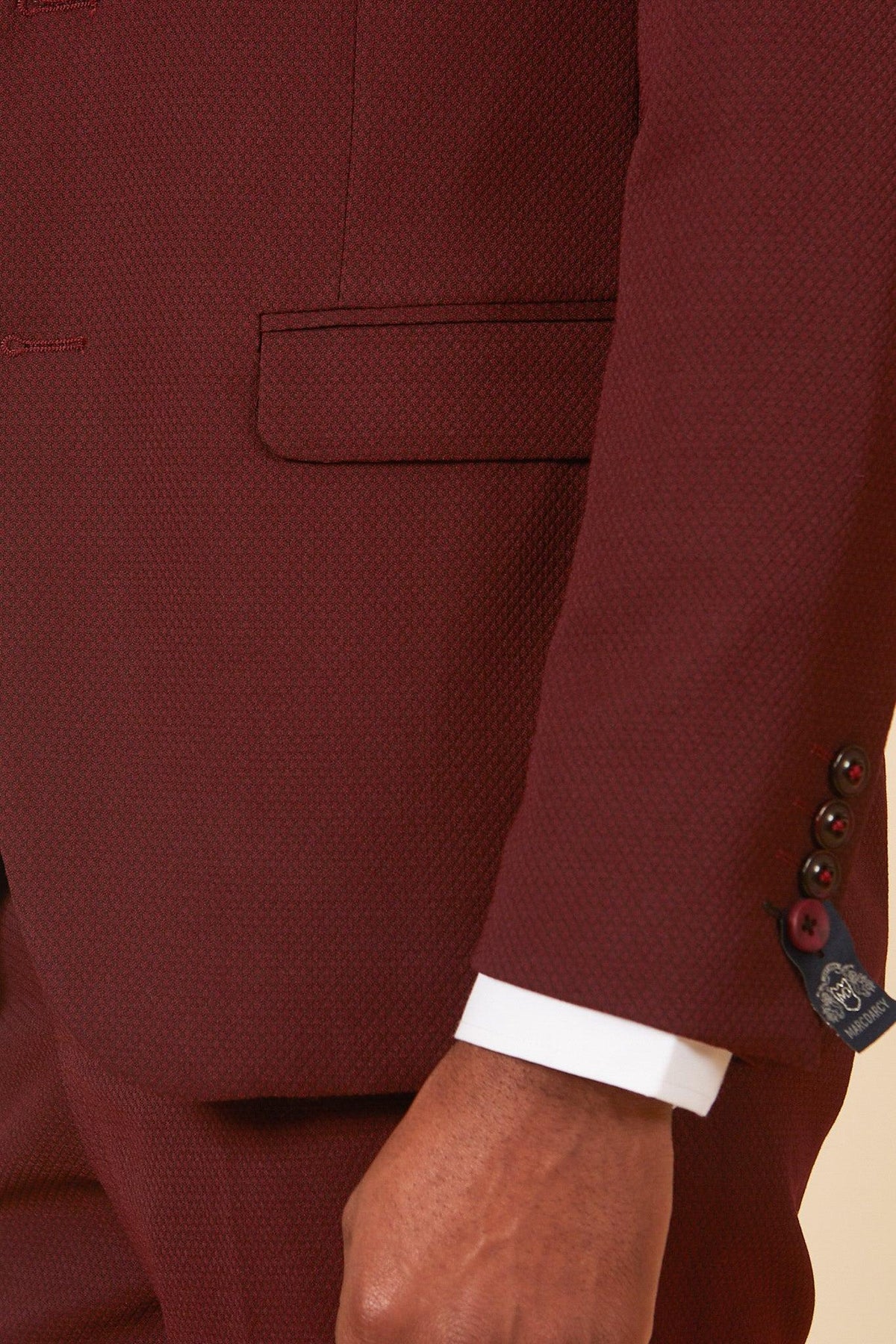 A close-up of a person wearing the marcdarcy MAX Wine Three Piece Suit with flap pocket, white shirt cuff, and brown sleeve buttons. This stylish slim fit suit is perfect for wedding guests; the person's hand rests by their side.