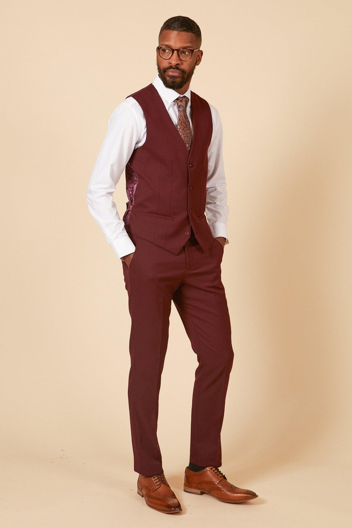 A man stands against a beige background wearing the marcdarcy MAX - Wine Three Piece Suit with a white shirt, patterned tie, brown shoes, and glasses. Left hand in his pocket, he looks right—ideal as a wedding guest suit.