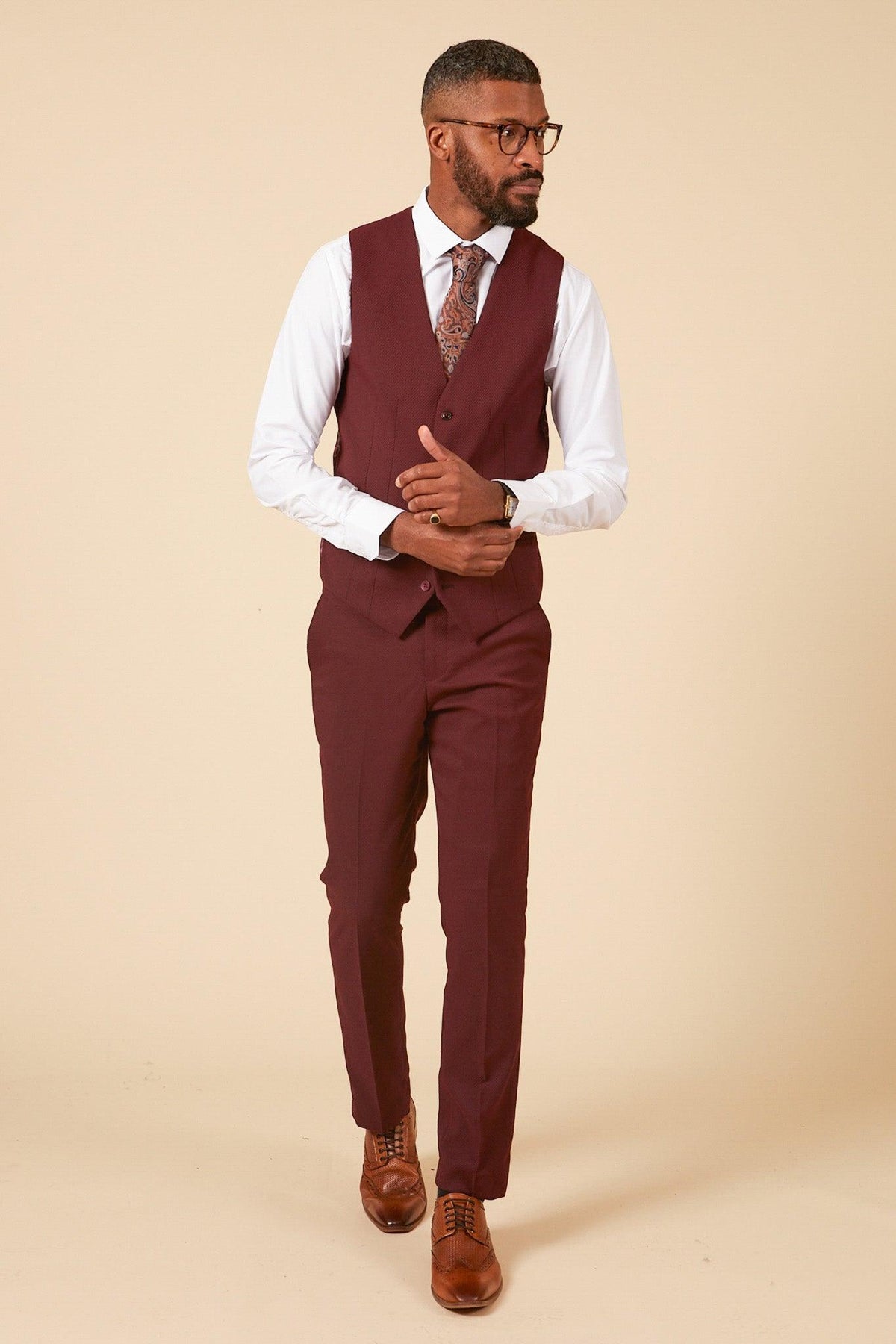 A man wearing glasses, a white dress shirt, the marcdarcy MAX - Wine Single Breasted Waistcoat with matching trousers, a patterned tie, and brown dress shoes stands against a beige background—a sharp wedding guest look.