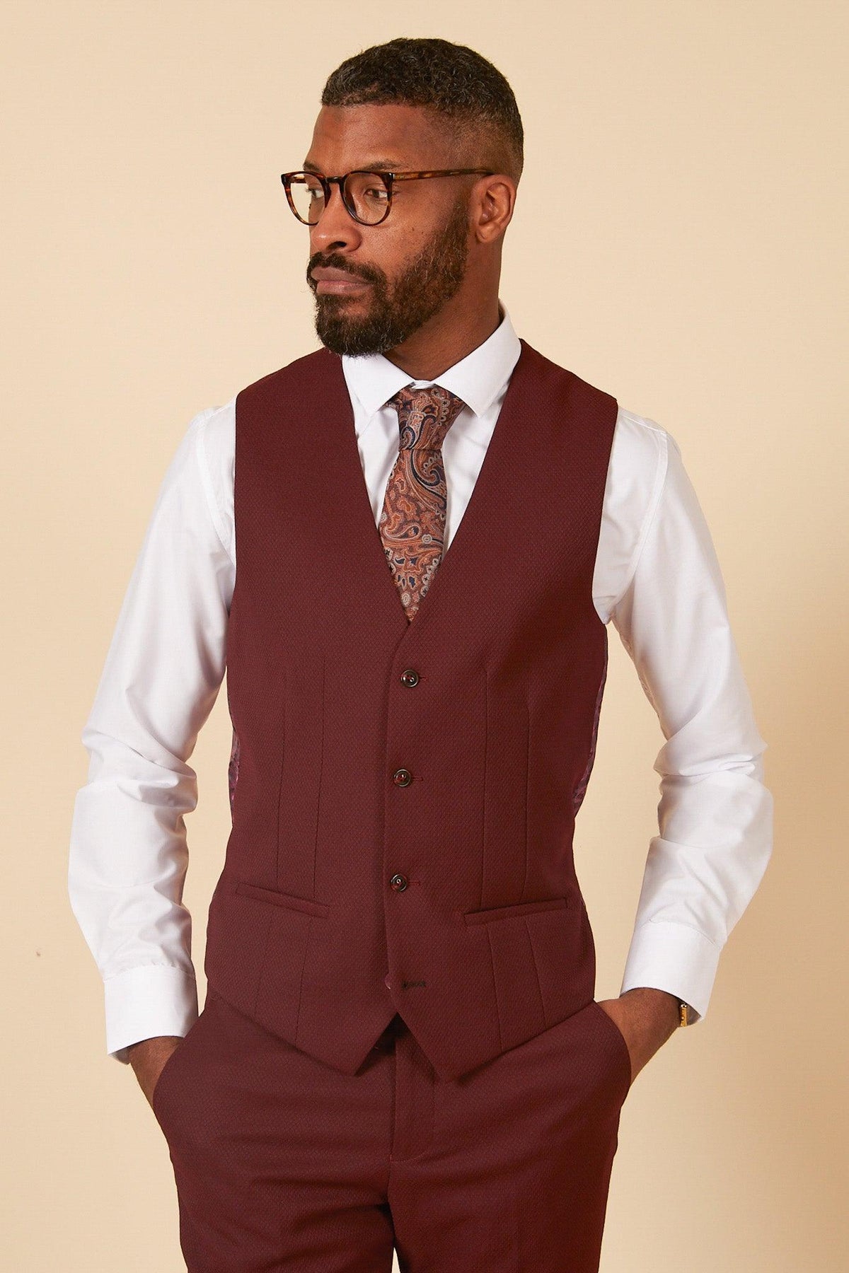 A man in glasses, a white dress shirt, patterned tie, and marcdarcy's MAX - Wine Single Breasted Waistcoat with matching pants stands hands-in-pockets against a beige background—a sharp wedding guest look.