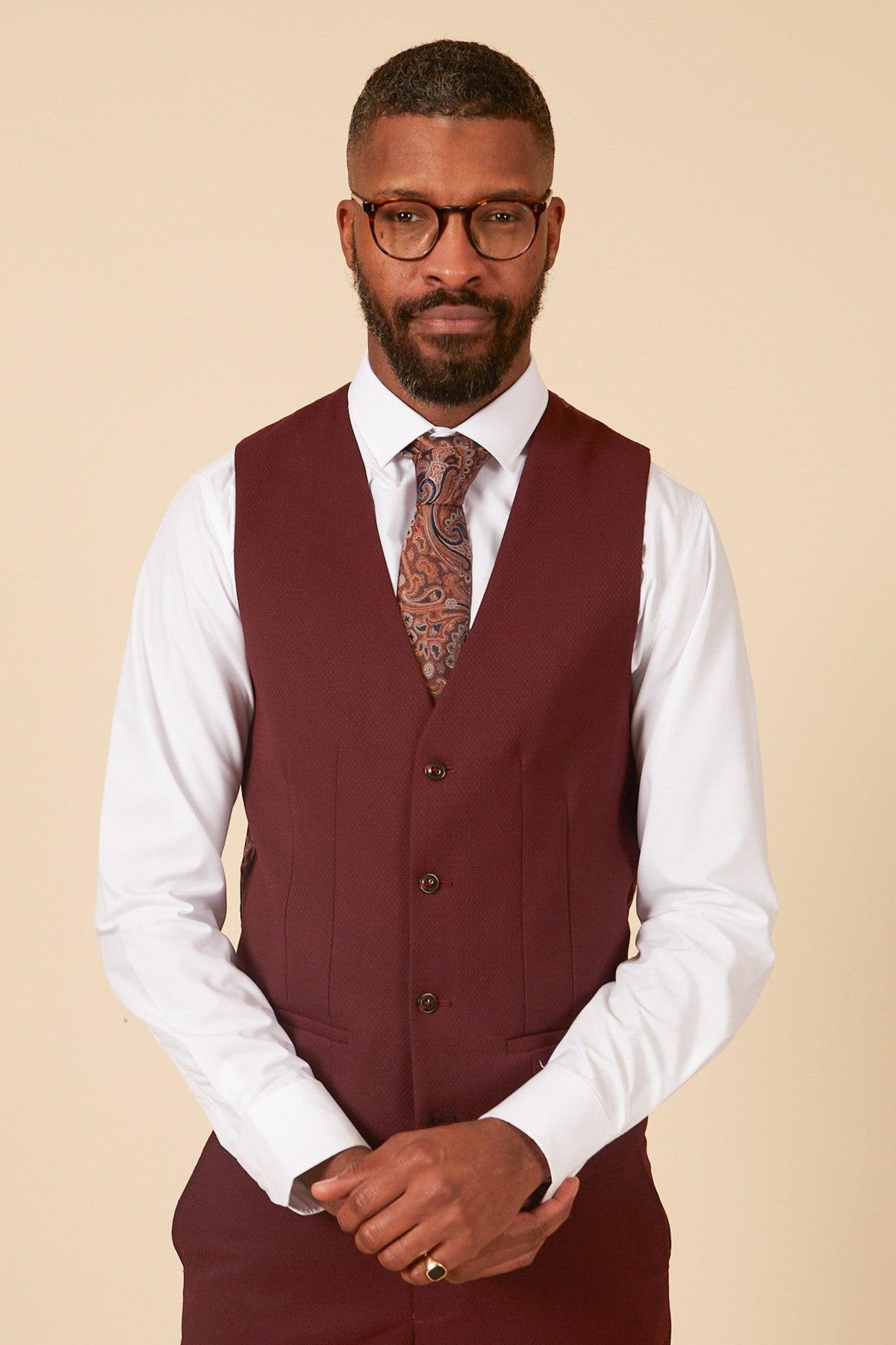A man with short hair, a beard, and glasses wears the marcdarcy MAX - Wine Three Piece Suit, featuring a white dress shirt and paisley tie. He stands against a beige background with his hands clasped in front of him.