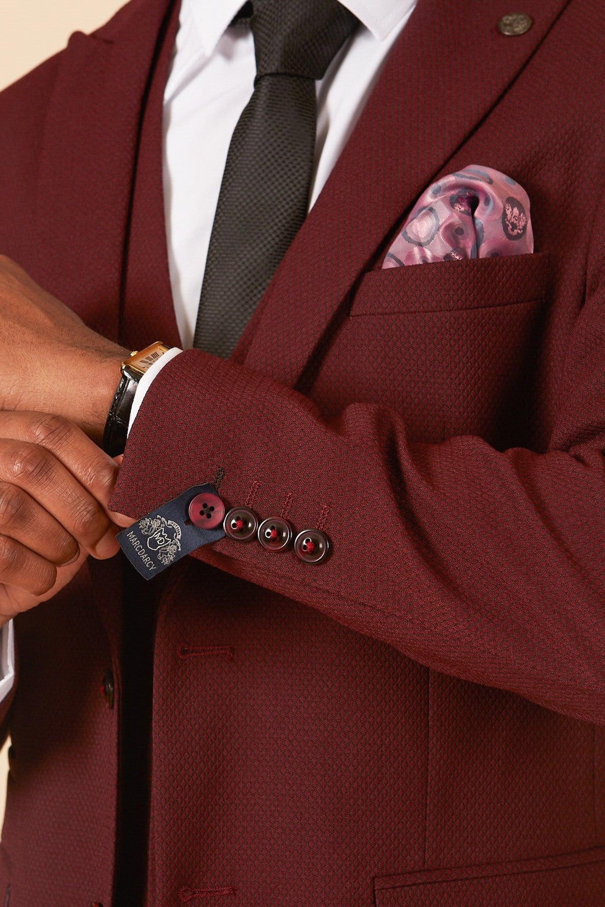 A person wears the marcdarcy MAX Wine Three Piece Suit with a black tie, four cuff buttons, and a pink patterned pocket square—an ideal, stylish look for a wedding guest.
