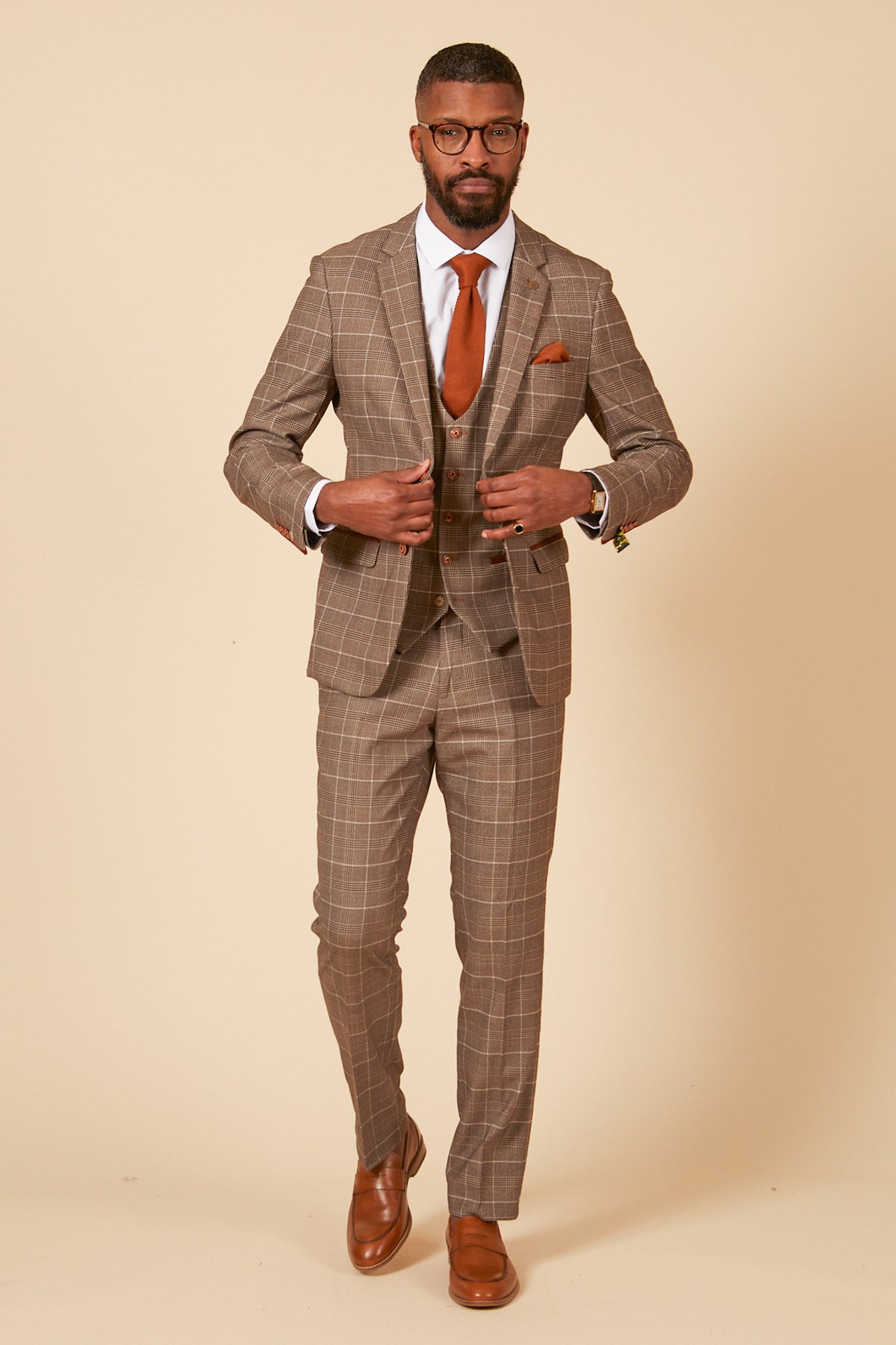 A Black man wearing the marcdarcy RAY - Tan Check Three Piece Suit styled with a slim fit blazer, brown shoes, white shirt, and an orange tie.