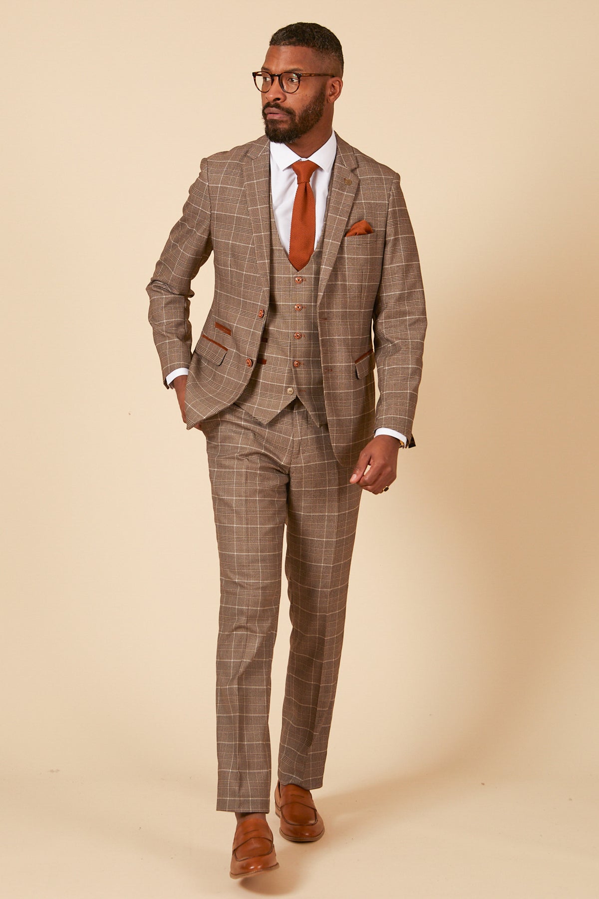 A man in a marcdarcy RAY Tan Check Three Piece Suit, paired with a white shirt, rust tie, pocket square, and brown shoes, stands confidently against a beige background—ideal for stylish office wear.