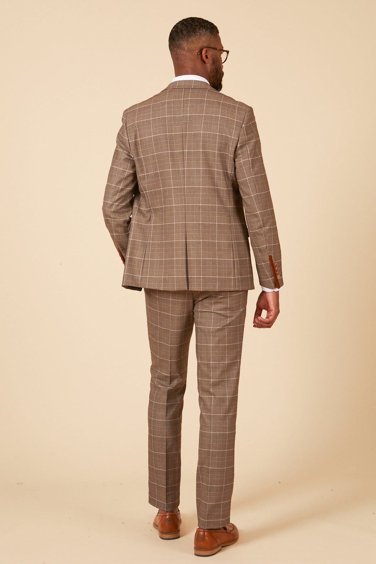 A man in the marcdarcy RAY Tan Check Three Piece Suit, white dress shirt, and brown dress shoes stands facing away from the camera against a plain beige background.