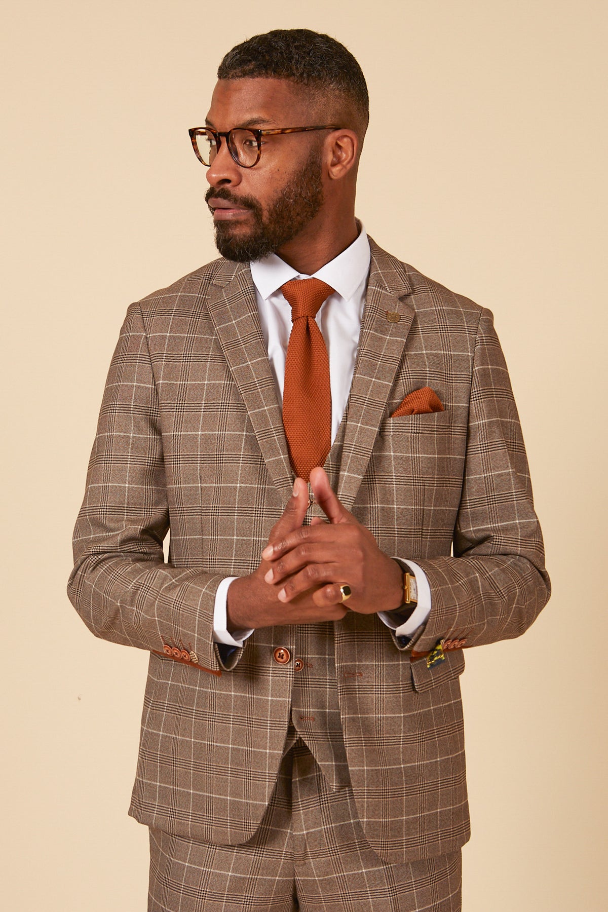 A man in the marcdarcy RAY Tan Check Three Piece Suit with a white shirt, rust tie, and matching pocket square stands against a beige background, hands clasped—a standout look for stylish office wear.