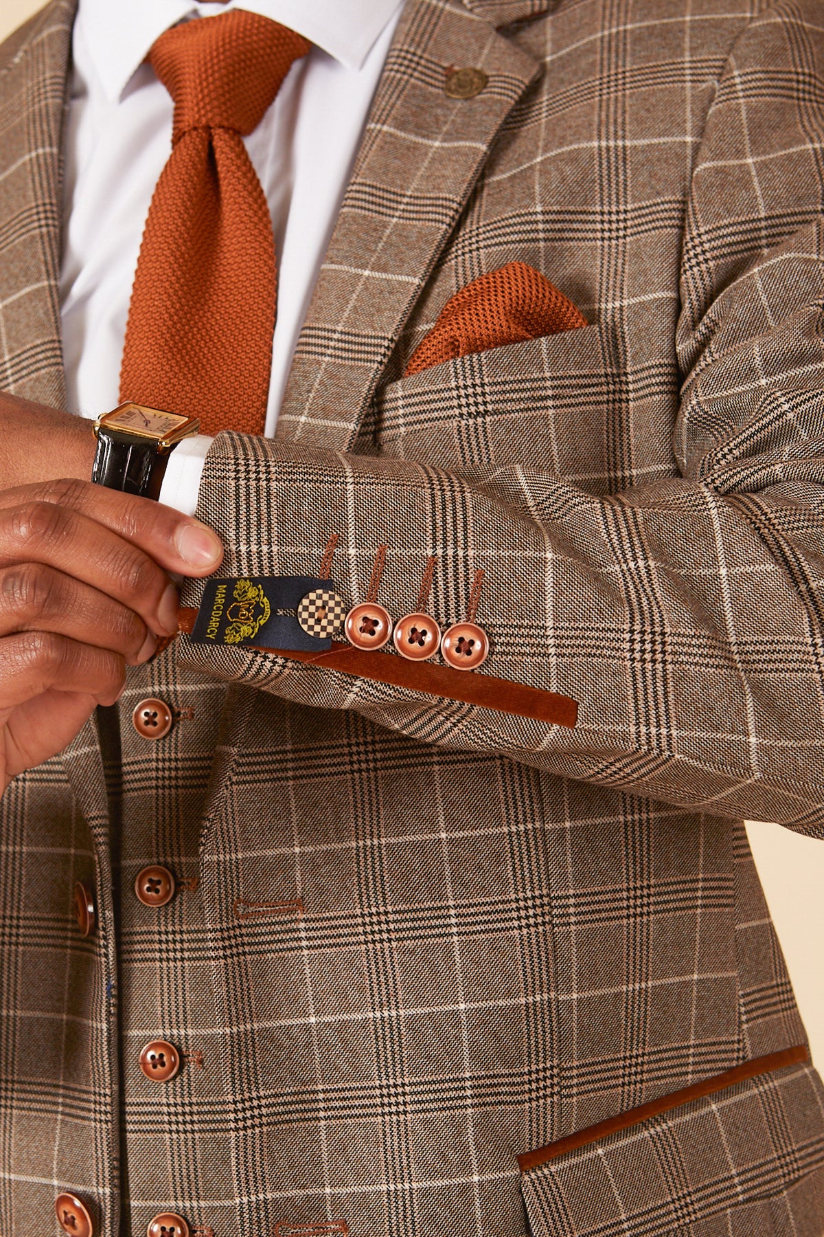 A person in the marcdarcy RAY Tan Check Three Piece Suit, styled with an orange tie and pocket square, adjusts their sleeve to reveal decorative lining and brown buttons. A wristwatch adds a refined touch—perfect for formal events.