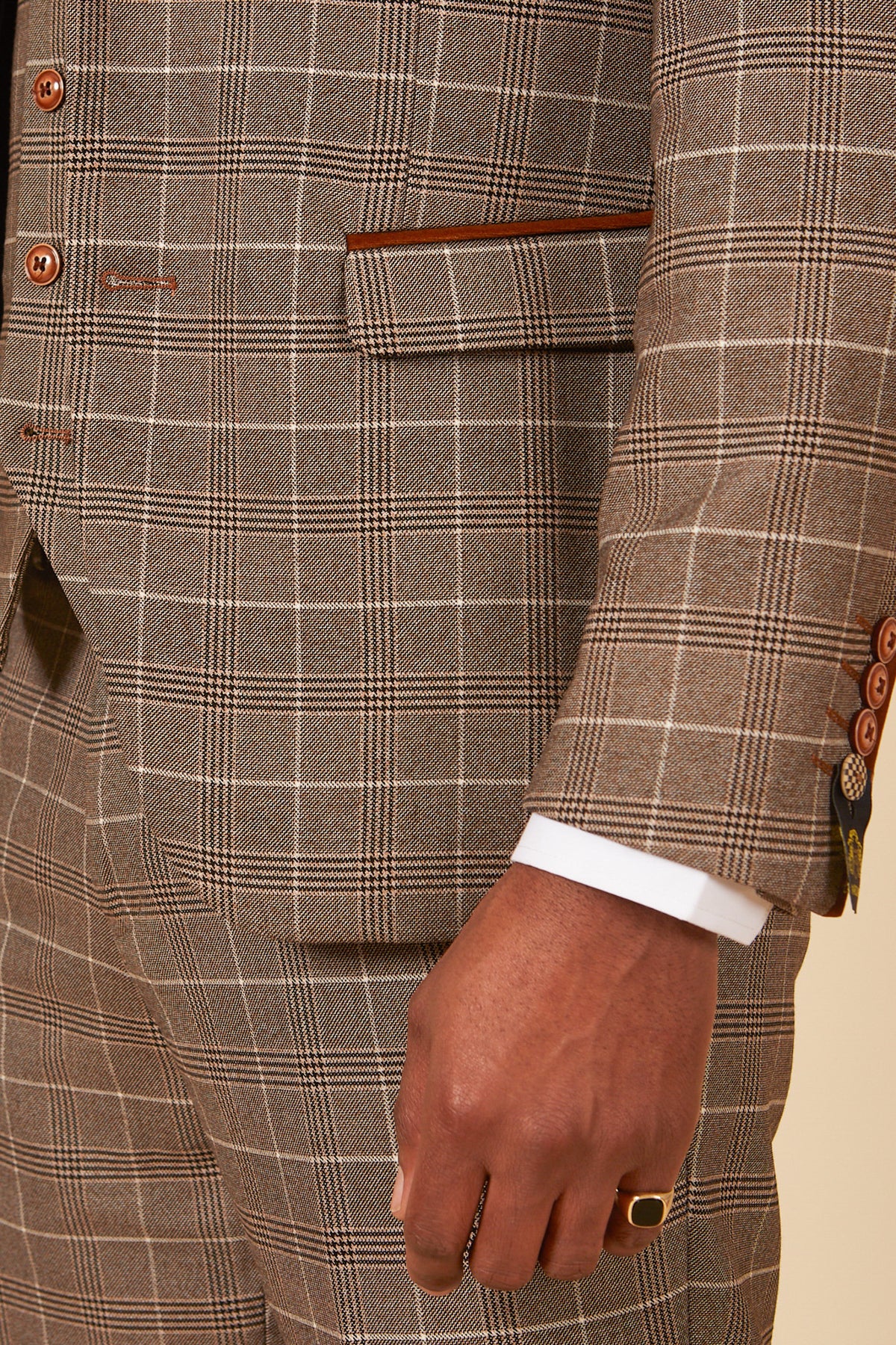 A close-up of a person wearing the marcdarcy RAY - Tan Check Blazer over a white shirt. Their right hand, with a gold ring, rests near the brown buttons and a pocket detailed with brown trim.