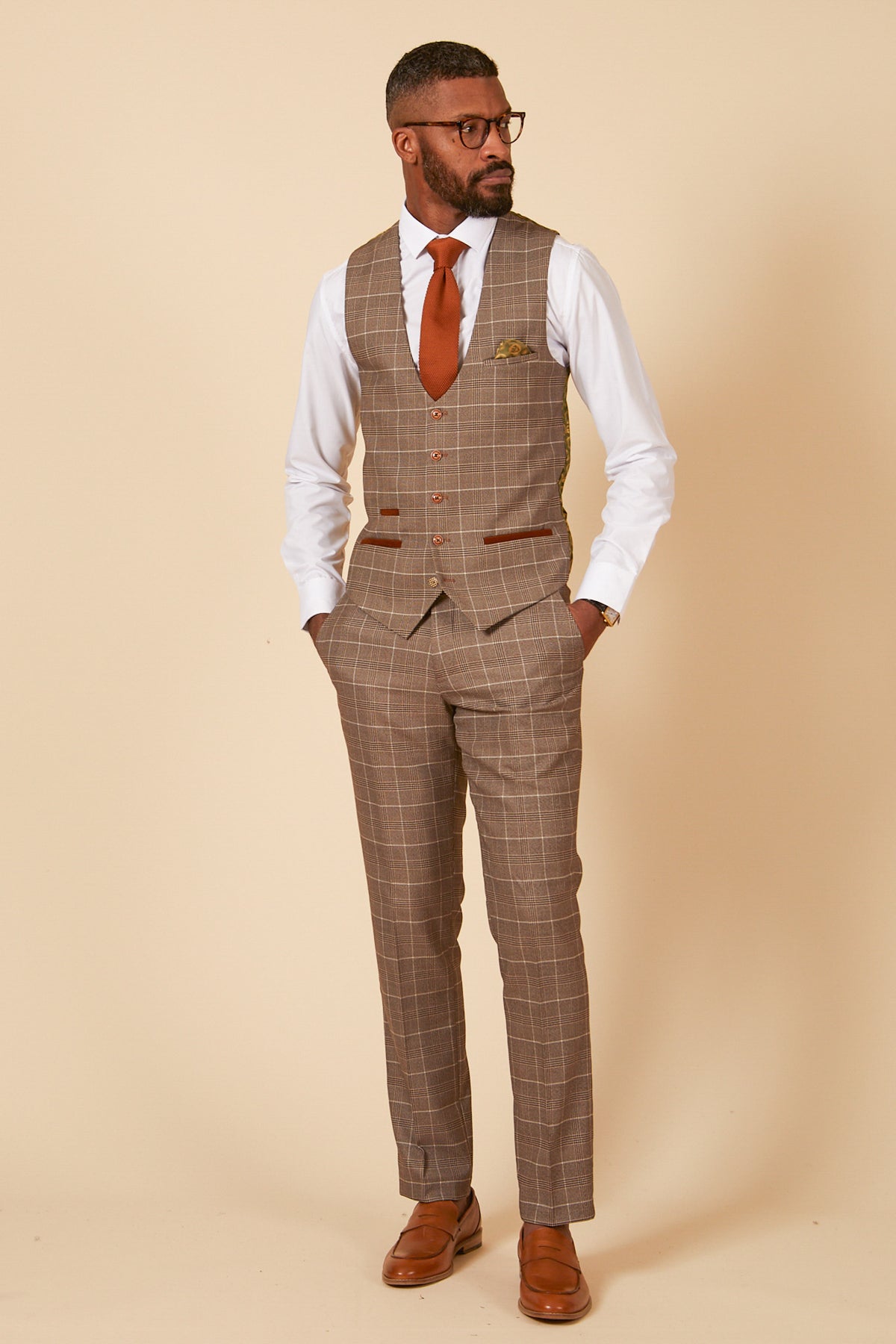 A man in glasses and formalwear—a marcdarcy RAY Tan Check Waistcoat with matching suit, white shirt, orange tie, and brown shoes—stands against a beige background, looking left with hands in his pockets.