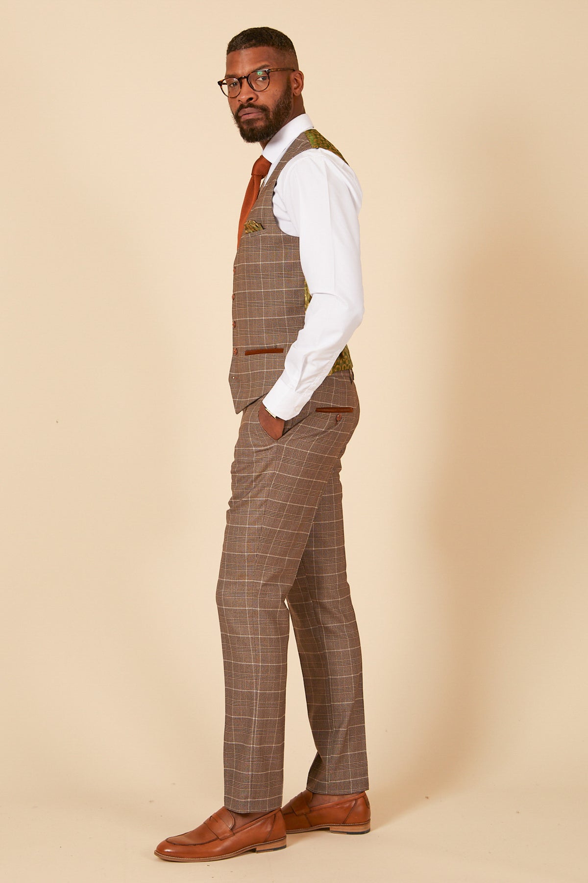 A man with a short beard stands sideways against a beige background, wearing glasses, a white shirt, tie, brown dress shoes, and the marcdarcy RAY - Tan Check Waistcoat with matching trousers. One hand is in his pocket.