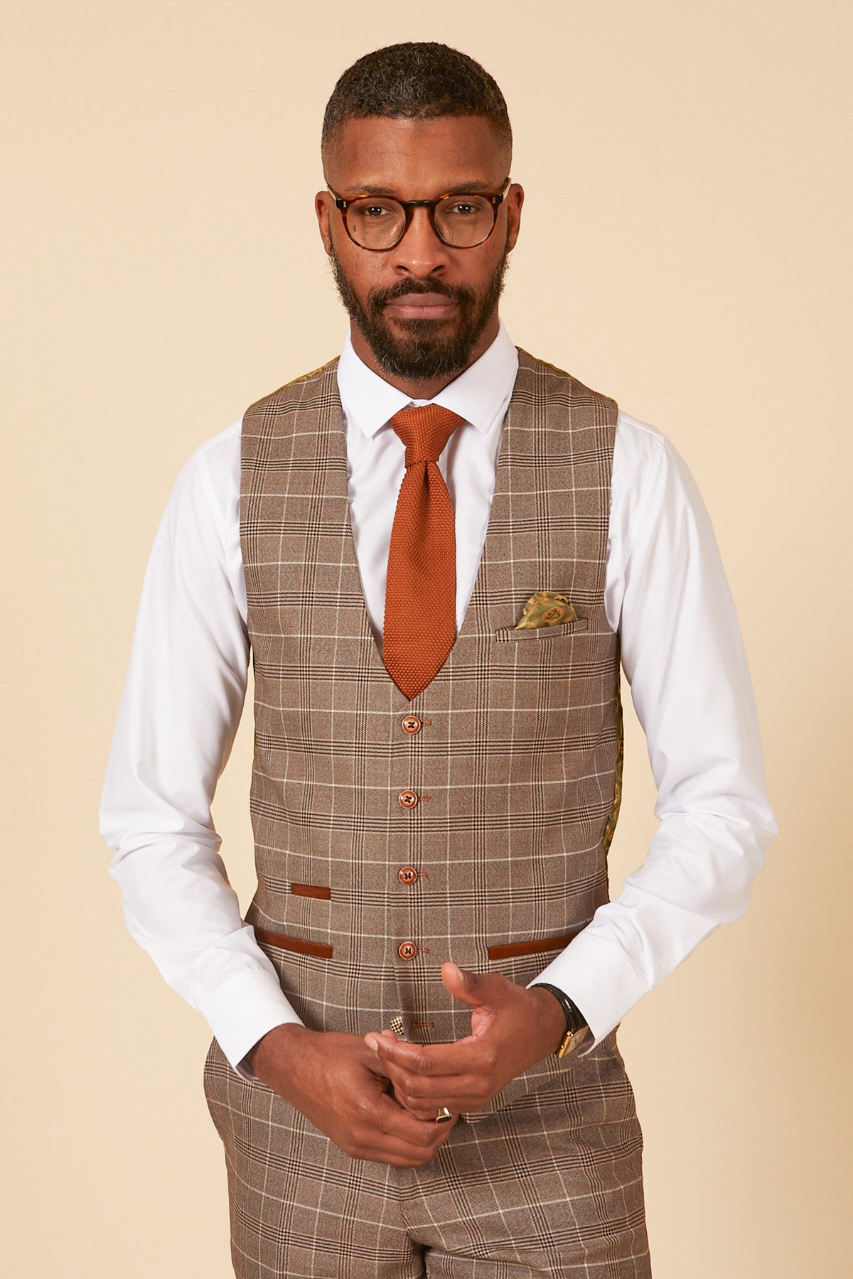 A man with short hair and a beard wears glasses, a white shirt, and the marcdarcy RAY Tan Check Waistcoat with a rust-colored tie. He stands against a plain beige background, looking directly at the camera.