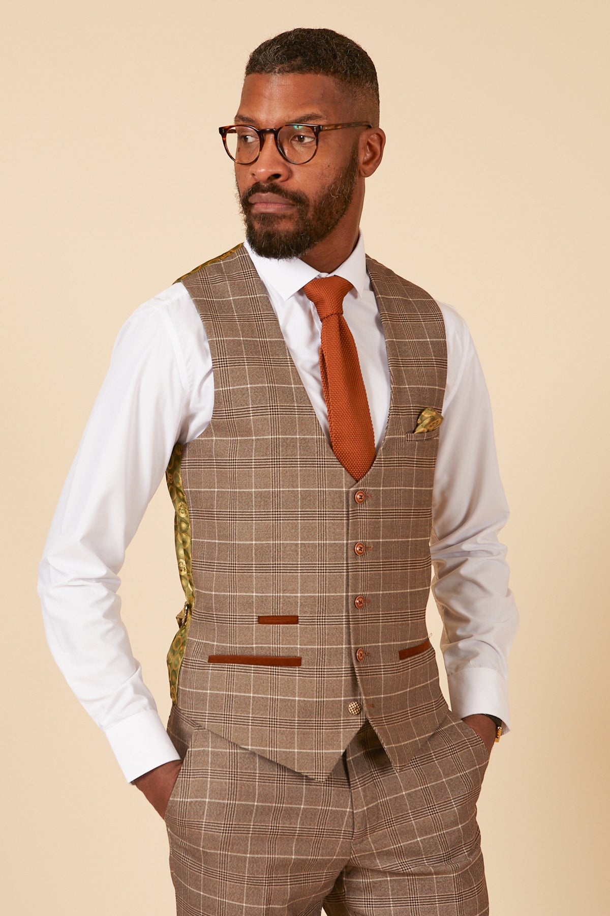 A man in glasses, a white dress shirt, and the marcdarcy RAY - Tan Check Waistcoat with matching pants and an orange tie stands with hands in pockets against a plain beige background.