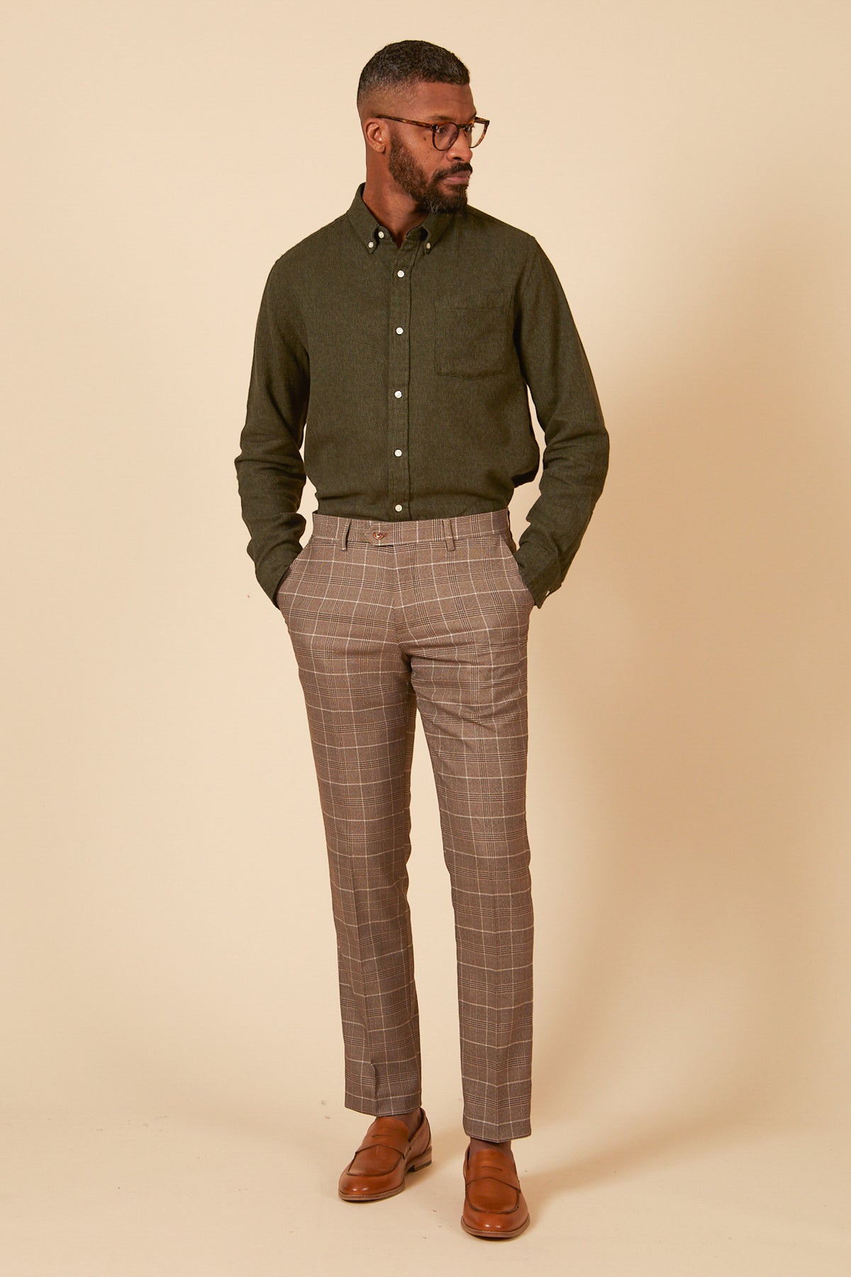 A man stands before a beige background, wearing glasses, a dark green shirt, marcdarcy RAY Tan Check Trousers, and light tan dress shoes, hands in his pockets and gazing to his left.
