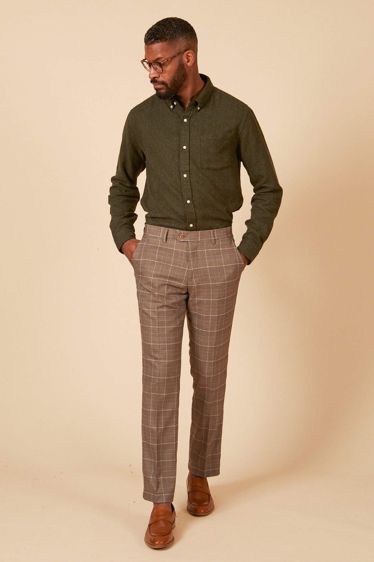 A man stands against a beige background wearing the marcdarcy RAY Tan Check Two Piece Suit, with his hands in his pockets, looking down and to the side.