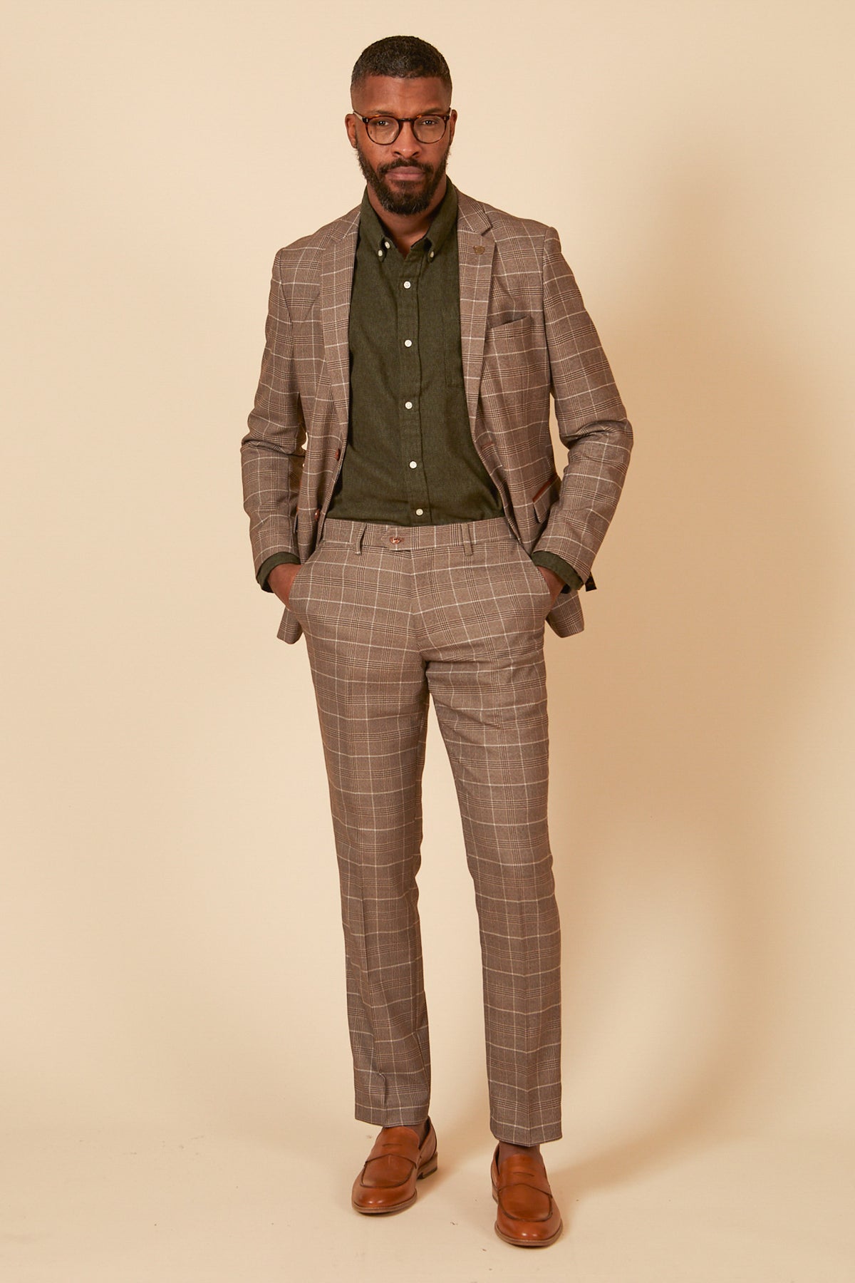 A man stands against a beige background wearing the marcdarcy RAY - Tan Check Blazer, an olive green button-up shirt, brown belt, brown shoes, and glasses. With hands in his pockets, he looks confidently at the camera.