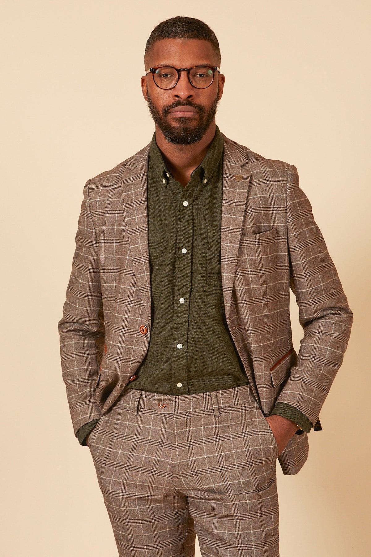 A man with short hair, a beard, and glasses wears the marcdarcy RAY - Tan Check Blazer over a dark green shirt. He stands against a beige background, hands in his pockets, looking confidently at the camera.
