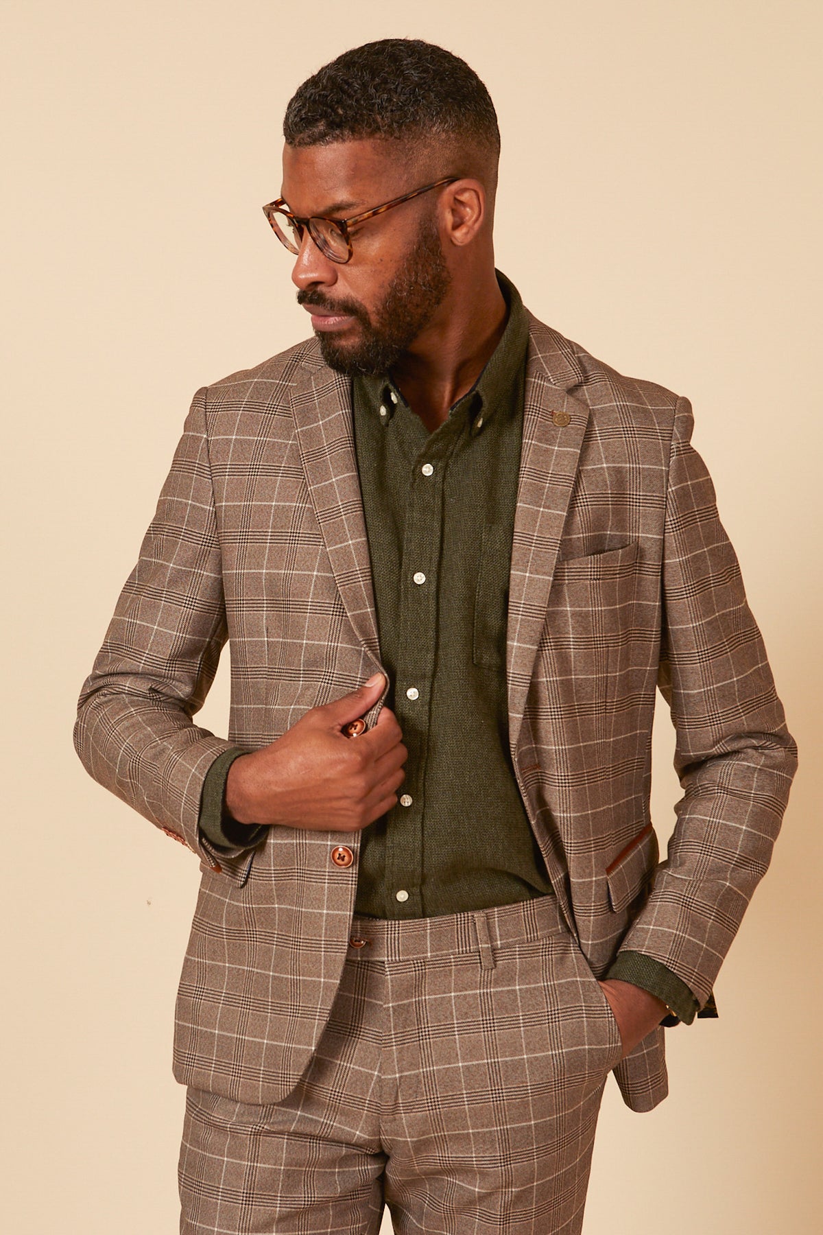 A man wearing glasses, the marcdarcy RAY - Tan Check Blazer, and a dark green shirt looks to his left, adjusting his jacket against a plain light beige background.