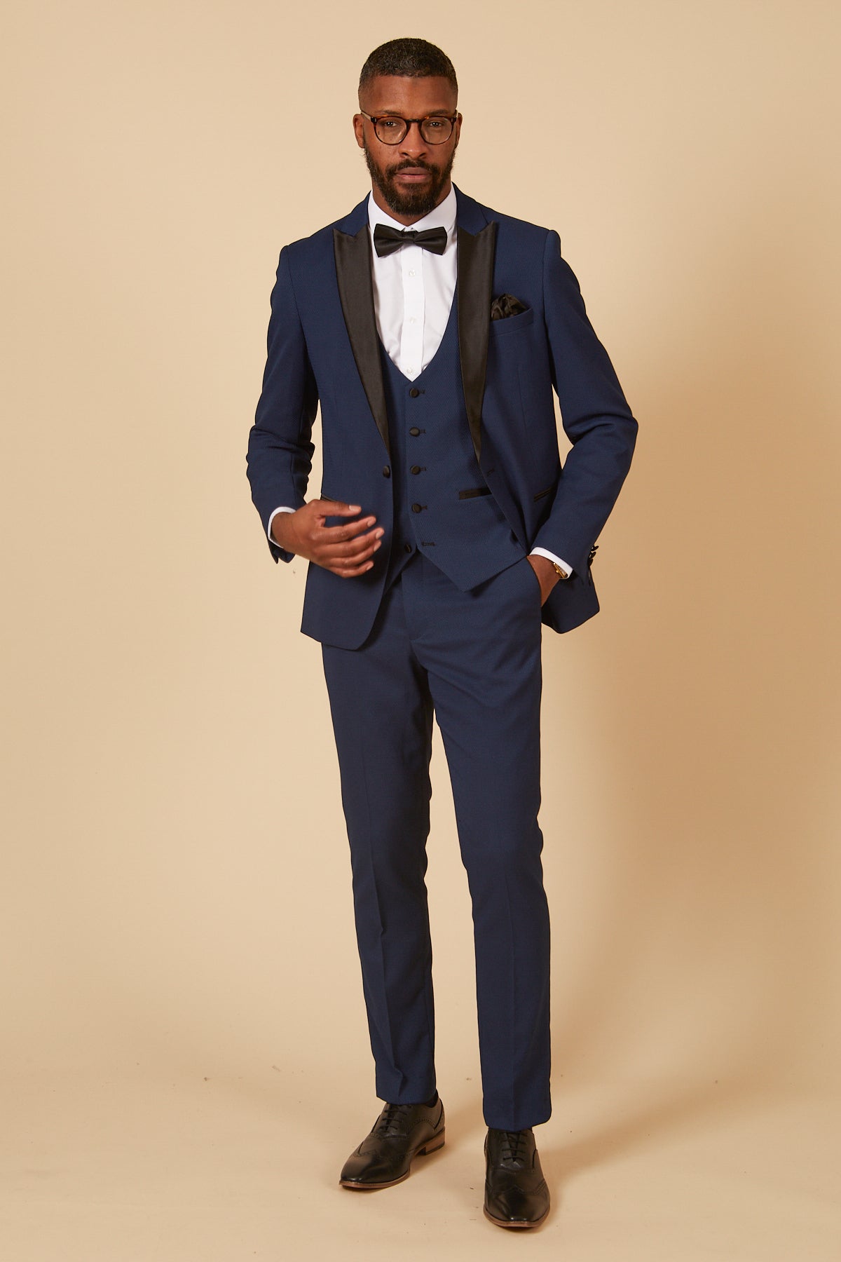 A man wears the marcdarcy Coronation Streets Liam Connor (Charlie Wrenshall) In DALTON Royal Blue Tux Suit, styled with a white shirt, black bow tie, glasses, pocket square, and black dress shoes against a beige background.