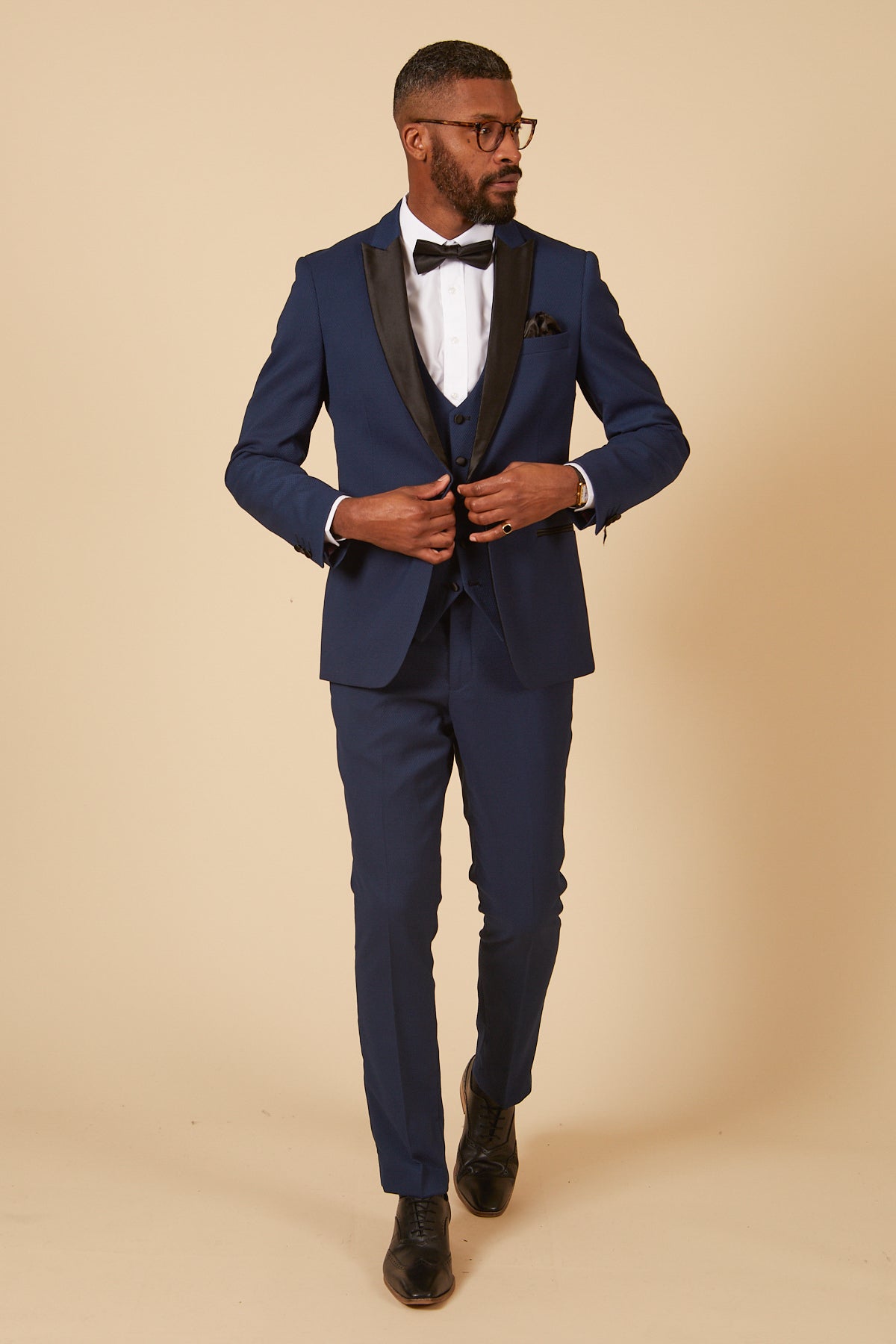 A Black man in a slim fit DALTON Royal Blue Tux Suit by marcdarcy, with black satin lapel, bow tie, white dress shirt, and black dress shoes adjusts his jacket buttons.