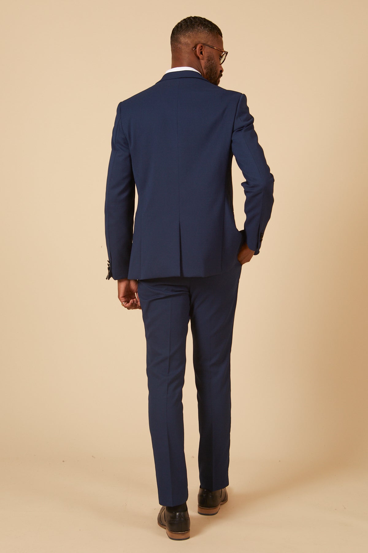 A man, dressed in a marcdarcy Coronation Streets Liam Connor (Charlie Wrenshall) in DALTON Royal Blue Tux Suit with a white shirt and black shoes, stands facing away from the camera against a beige background, one hand in his pocket.