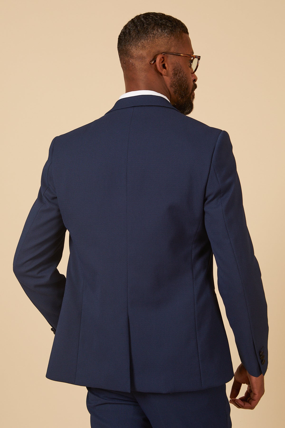 A man in marcdarcy’s DALTON Royal Blue Tux Lapel Diamond Blazer and glasses is shown from behind, standing against a beige background.